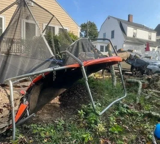 A damaged trampoline with its frame bent and fabric torn, lying on the ground in a backyard with houses and a car in the background.