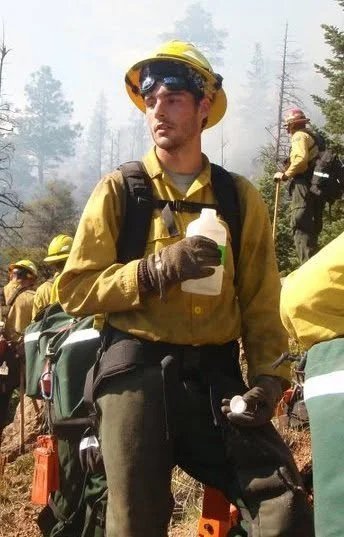A firefighter in yellow gear and helmet holding a spray bottle, with others working in a smoky forested area in the background.