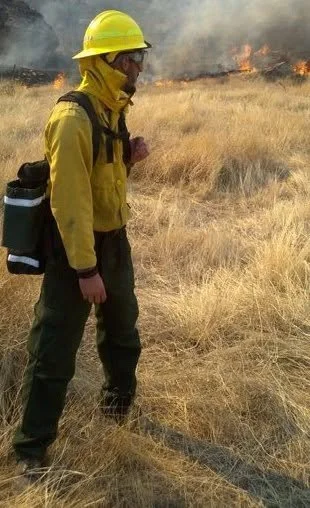 Firefighter in yellow gear and helmet monitoring a wildfire in a grassy field.