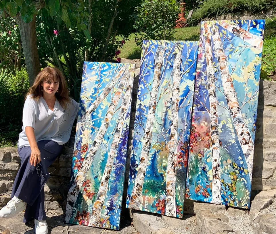 A woman sitting outdoors next to four large colorful paintings of birch trees with blue sky backgrounds.