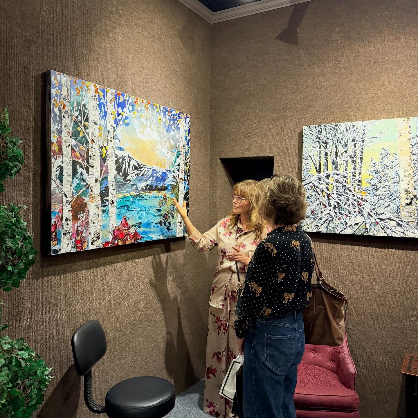 Two women are talking and looking at a colorful landscape painting in an art gallery. One woman is wearing a floral dress and glasses, explaining the artwork, while the other woman wears a black blouse with a tiger pattern and jeans.