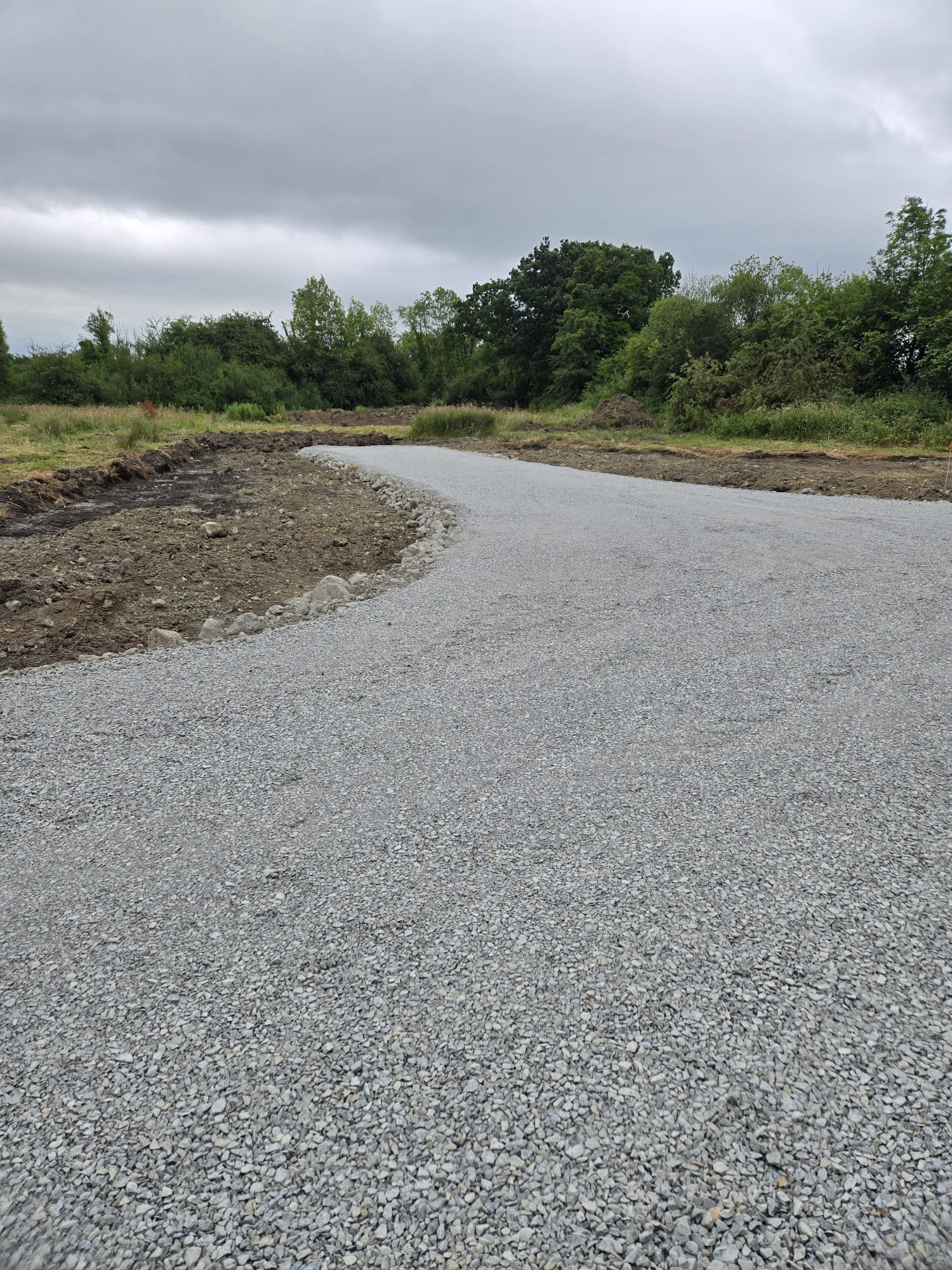 A gravel road curves to the left in a rural area, with lush green trees and bushes on either side, and a cloudy sky overhead.
