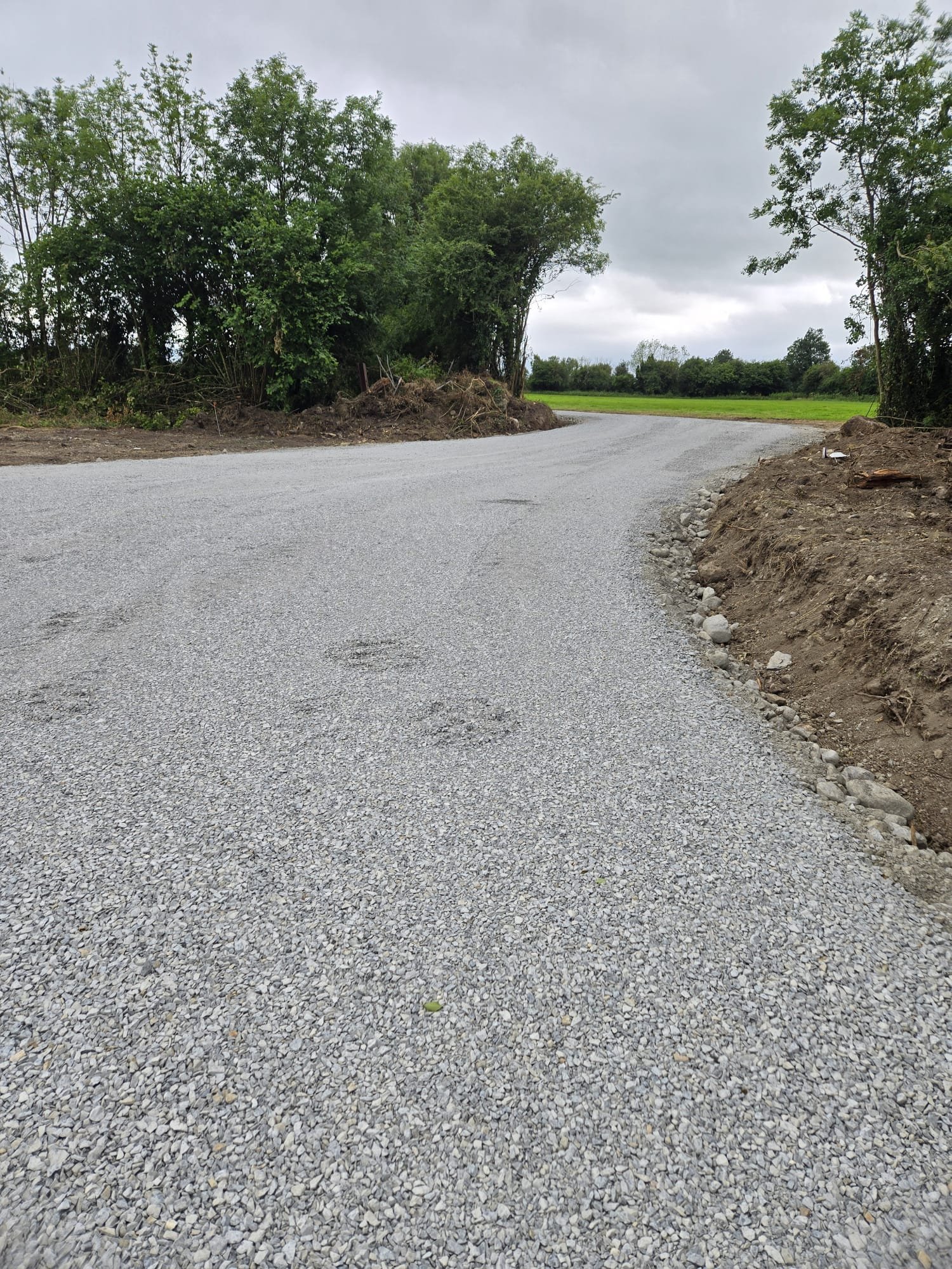 A gravel road curves to the right, flanked by dirt and small rocks on the edges. Dense green trees line the left side, and a field with more trees is visible in the background. The sky is overcast with gray clouds.