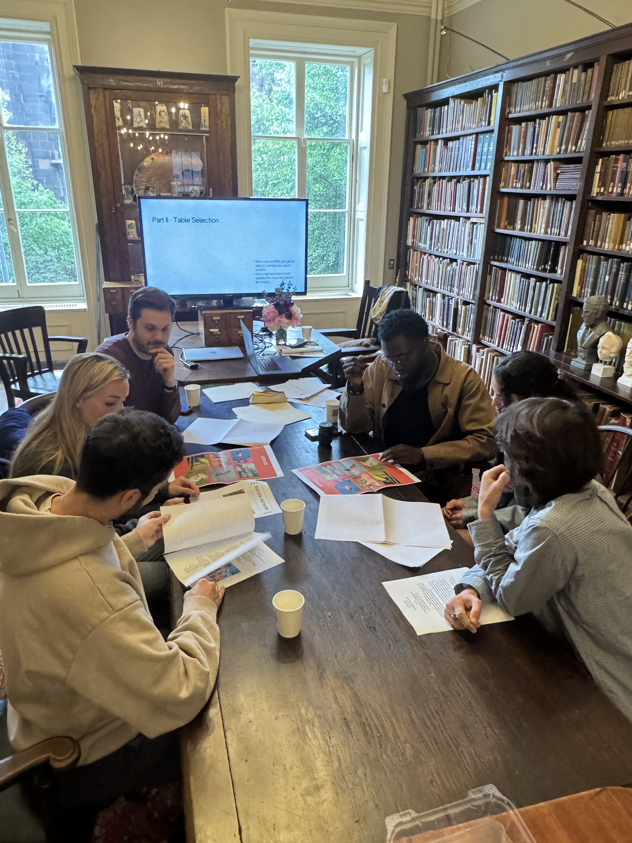 A group of people sitting around a large wooden table in a room with tall windows and bookshelves, engaged in a meeting or discussion, with papers and magazines on the table and a large screen displaying a presentation.
