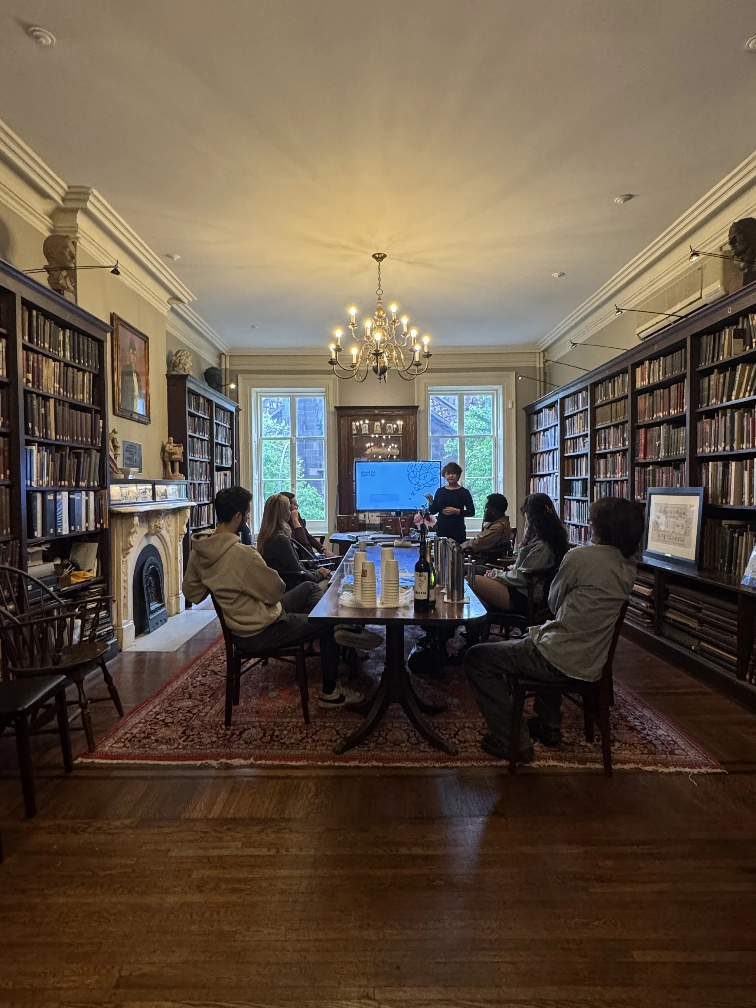 A group of people participating in a discussion in a library or study room with wooden bookshelves, large windows, and a chandelier, while a woman stands at the front near a digital display.