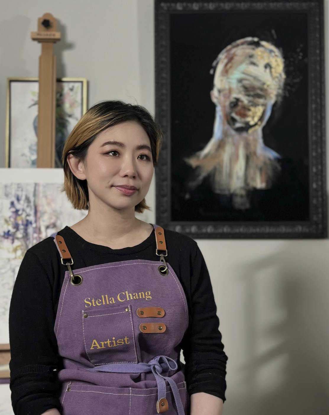 A woman with short hair wearing a black shirt and a purple apron that has her name 'Stella Chang' and the word 'Artist' on it, standing in an art gallery. Behind her, there are various artworks including a large abstract portrait with a blurred face on the wall.