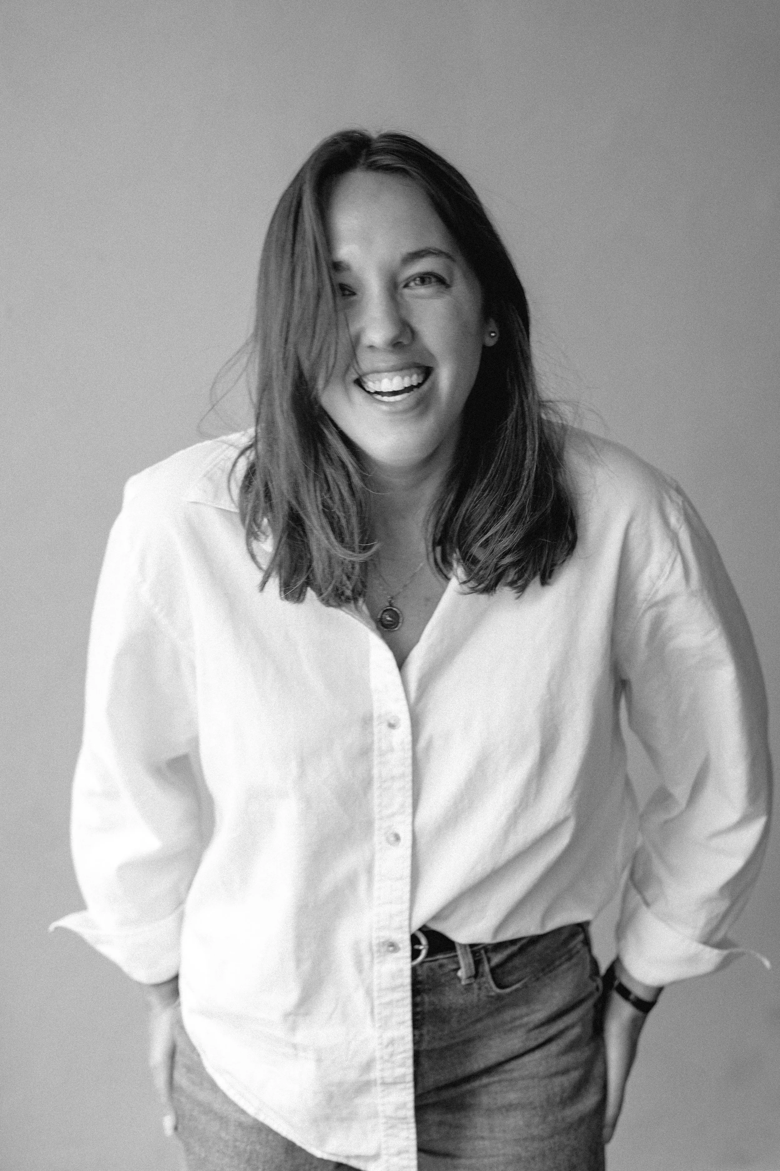A woman with medium-length hair, smiling and wearing a light-colored shirt and jeans, standing against a plain wall.