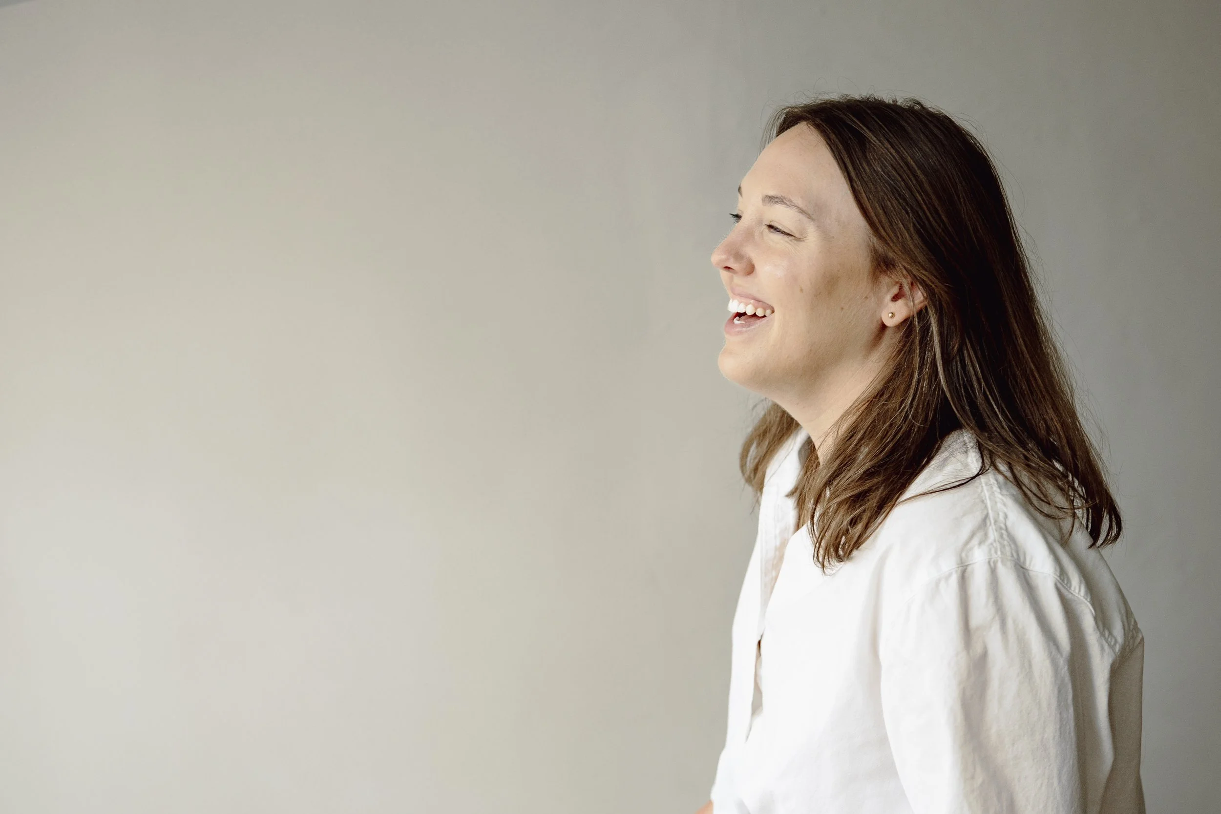 Profile of a woman with brown hair smiling and looking slightly to the side, wearing a white shirt, against a plain neutral background.