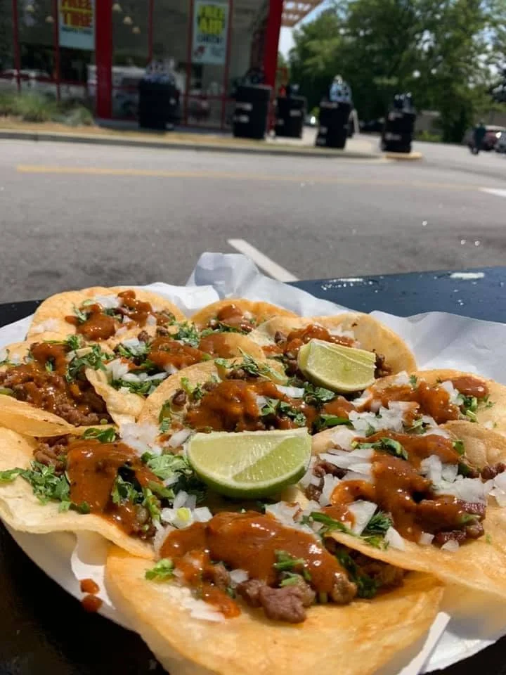 Close-up of three beef tacos on a plate, topped with chopped onions, cilantro, hot sauce, and lime wedges, with a parking lot and trees in the background.