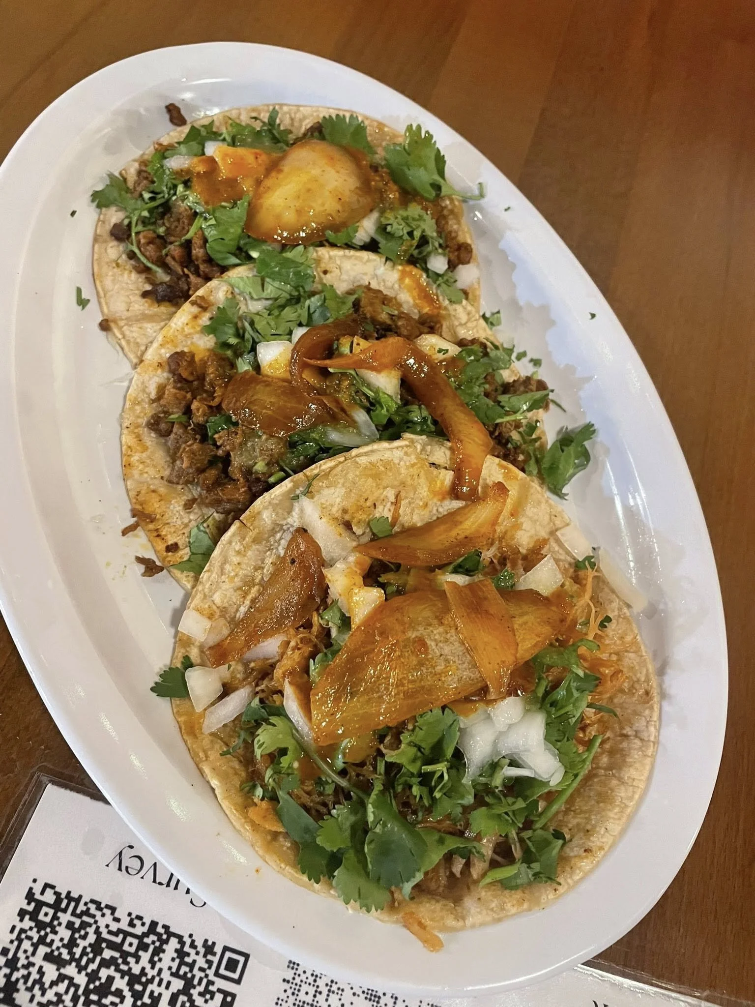 Three tacos topped with chopped onions, cilantro, and some sauce on a white oval plate.