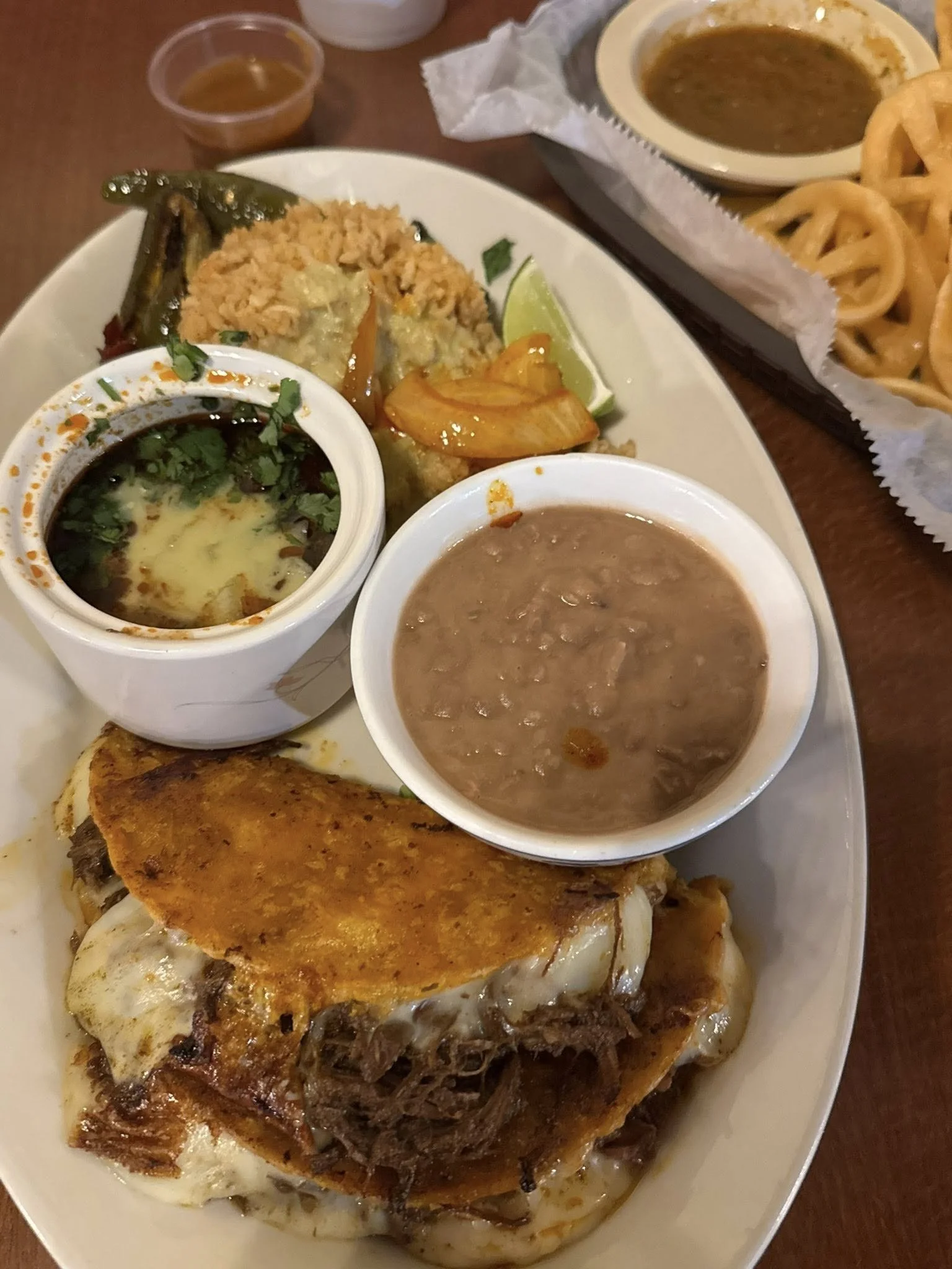 Plate of Mexican food including a large bowl of refried beans, a small bowl of salsa, a tortilla with fajita beef, rice, fried onions, lemon wedges, and a fried steak with eggs, alongside a basket of fried onion rings.