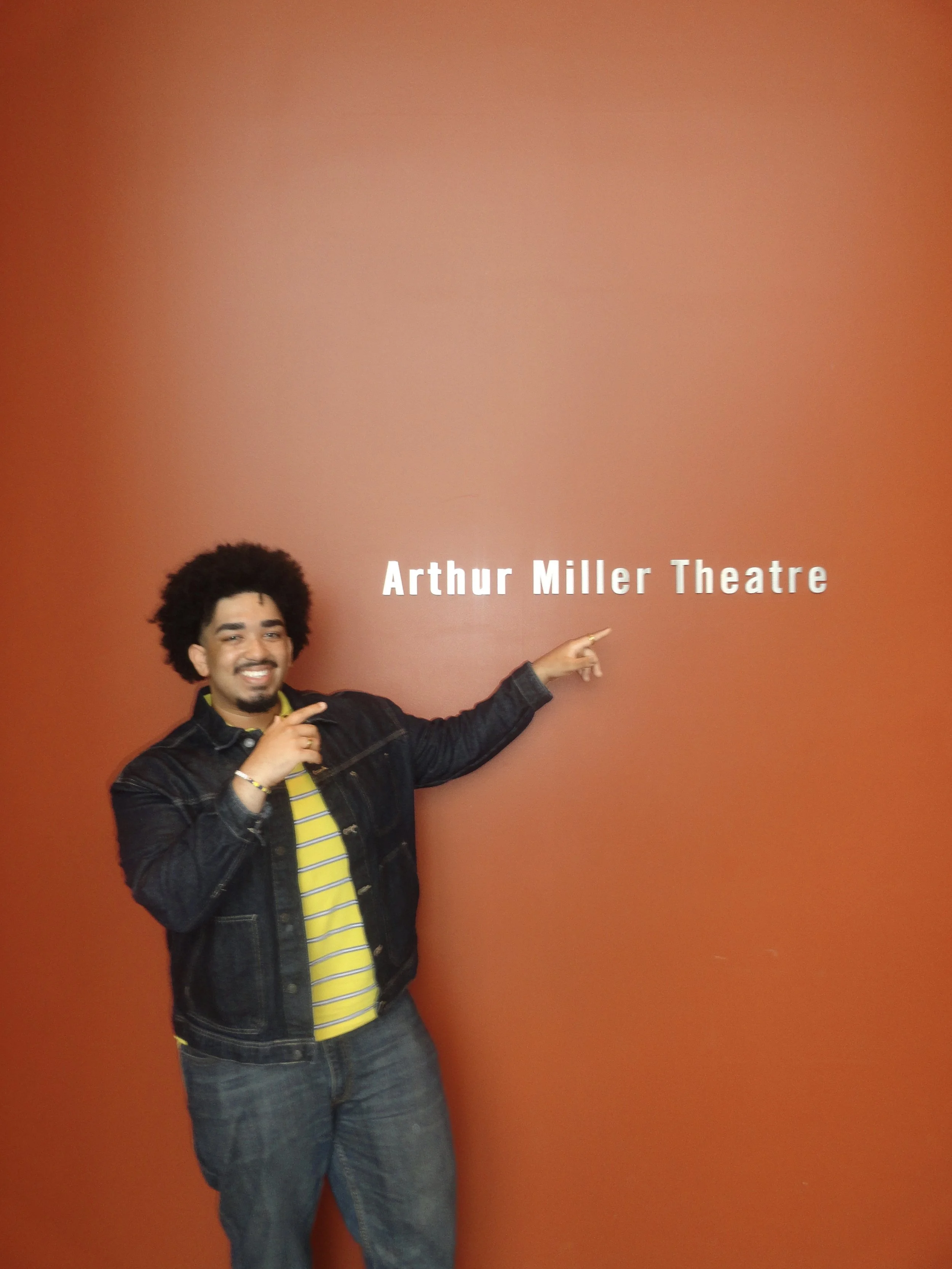 A man with curly hair and a goatee smiling and pointing at a sign that reads "Arthur Miller Theatre" against a reddish-orange wall.