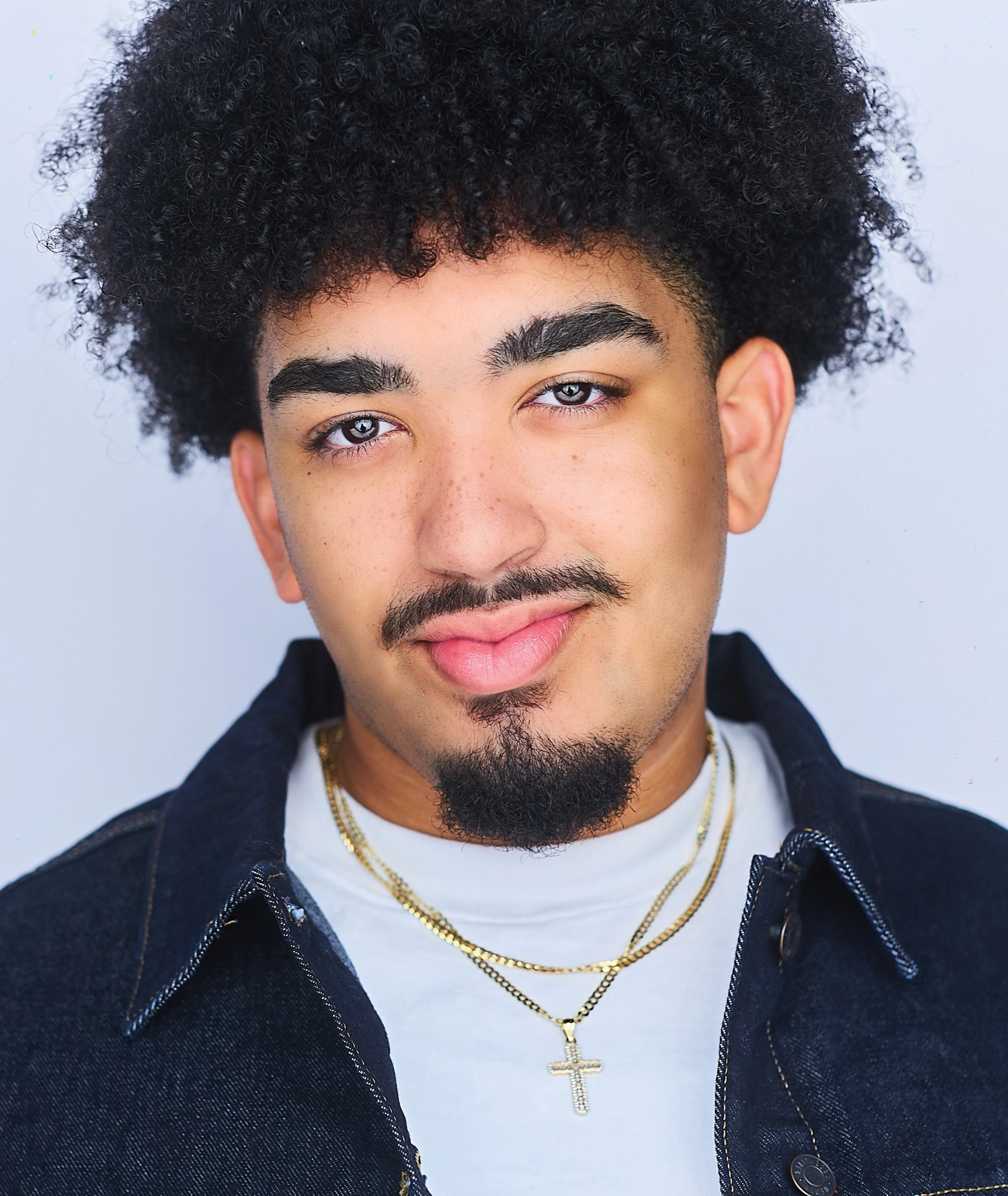 Close-up of a young man with curly black hair, light skin, and gray eyes, wearing gold necklaces, a white shirt, and a denim jacket, against a plain light background.