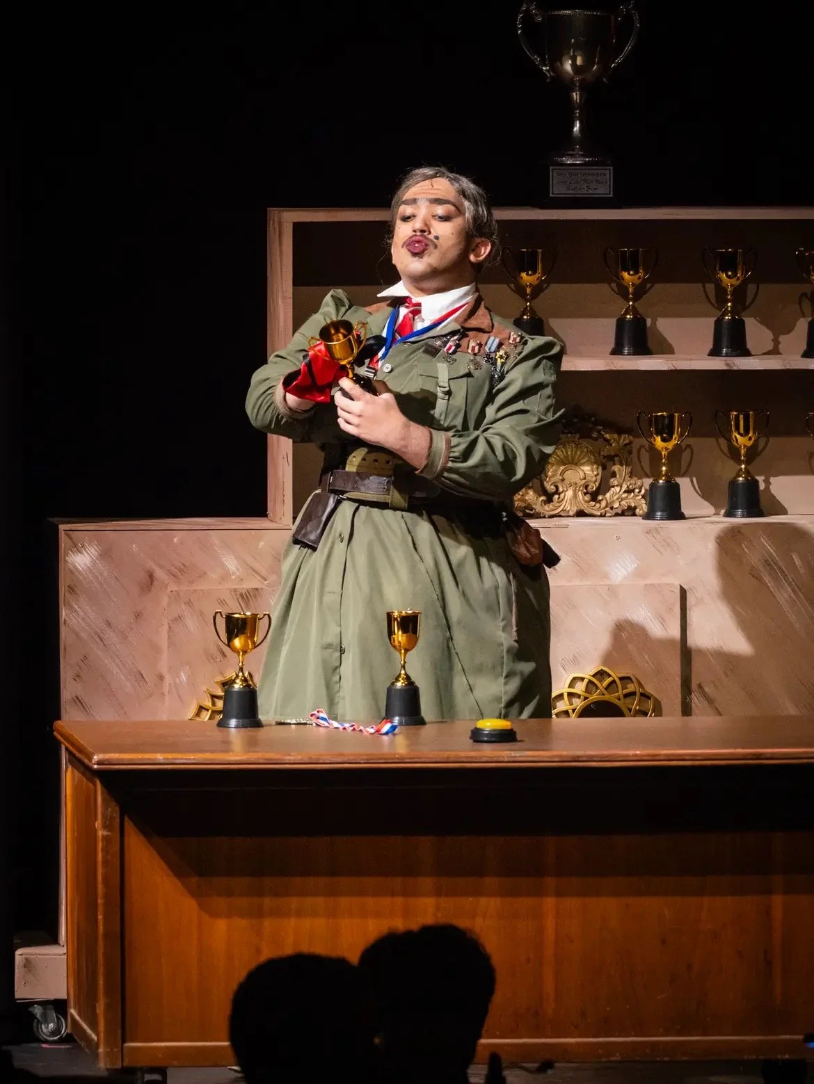 Person dressed in military costume holding a trophy, standing at a table with more trophies and medals, in a setting that appears to be an awards ceremony or performance.