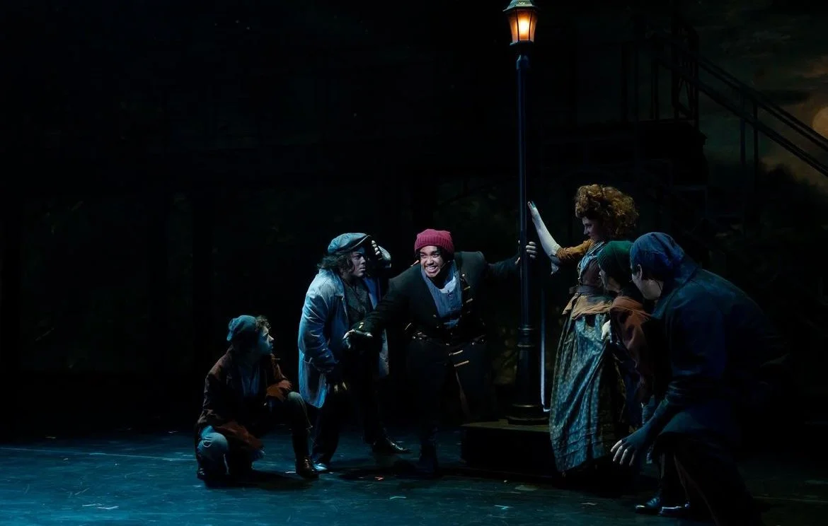 Theater performance of six actors on stage, with dim lighting, resembling a scene from a play set outdoors at night.