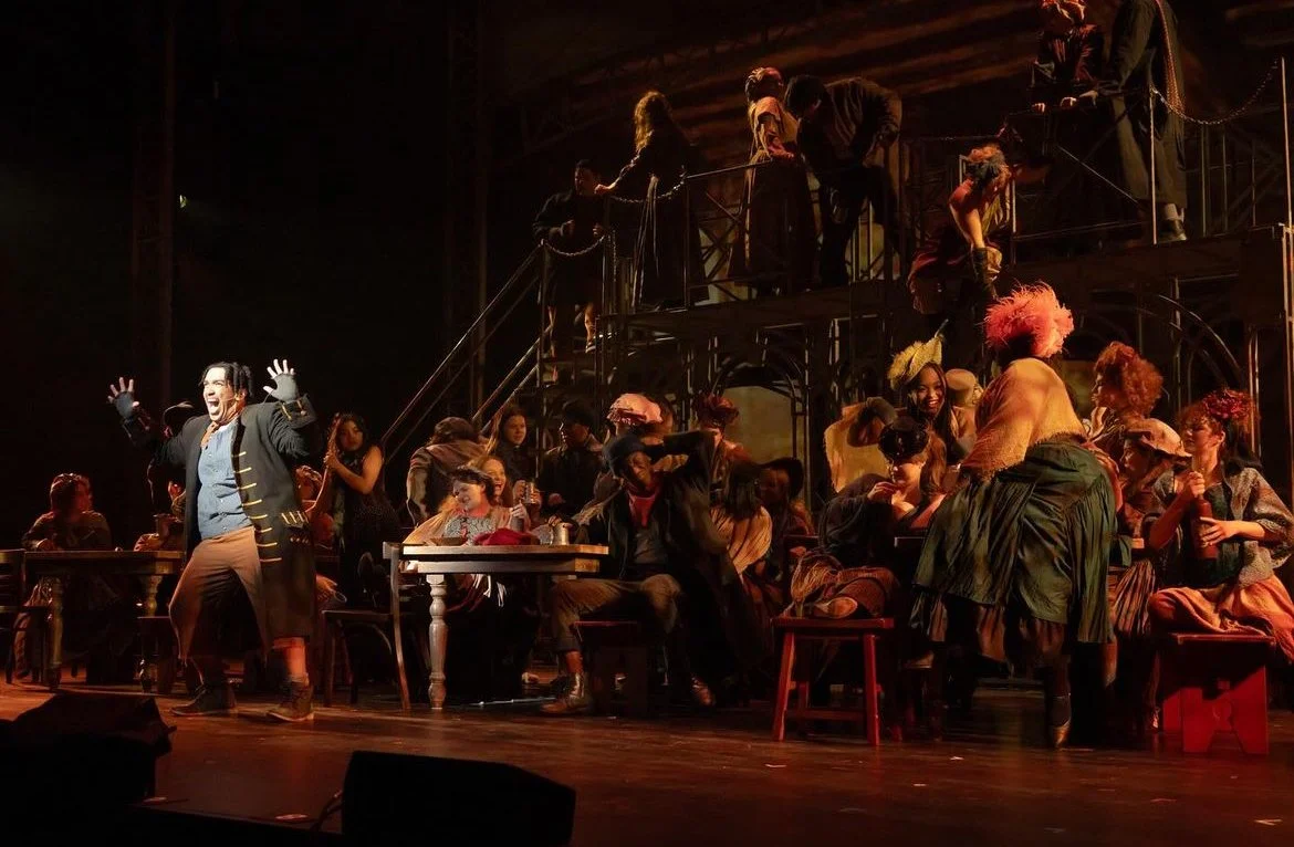 Theater cast performing on stage with many actors in period costumes, some sitting and some standing, with a two-level set and dim lighting.