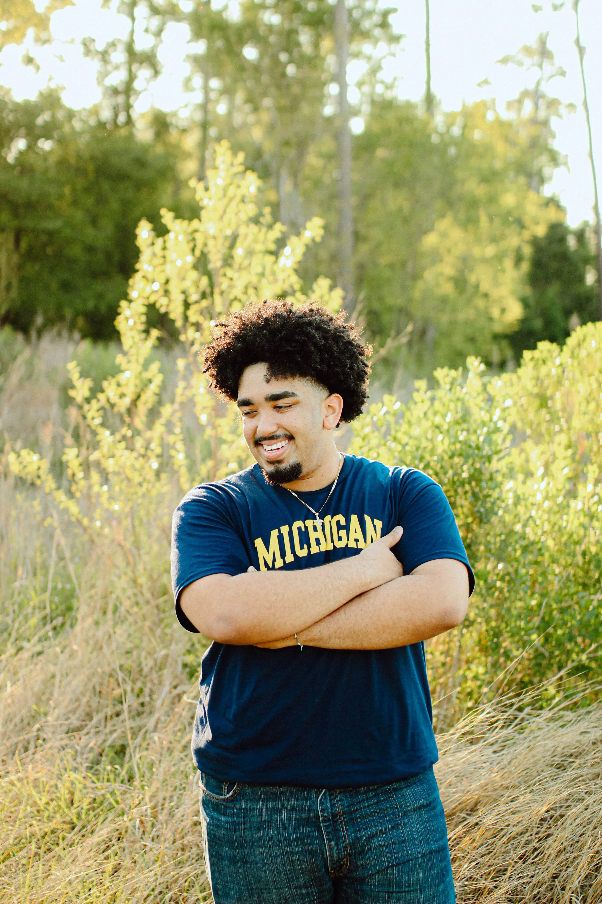 A young man with curly hair and a beard smiling outdoors in a grassy field with yellow flowers, wearing a navy blue Michigan t-shirt and crossed arms.