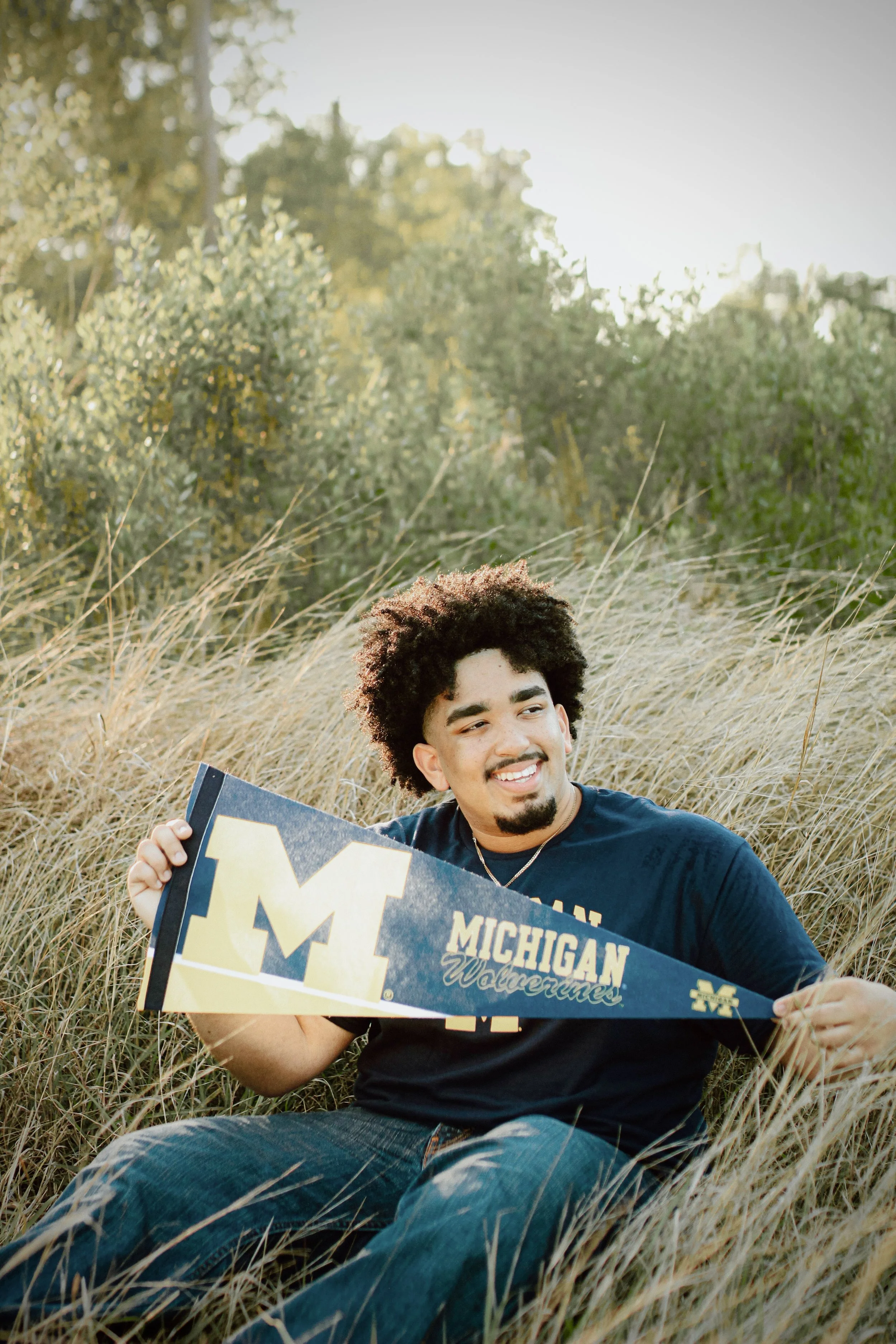 A young man sitting in tall grass outdoors, holding a Michigan Wolverines banner and smiling. The background features trees and a clear sky.