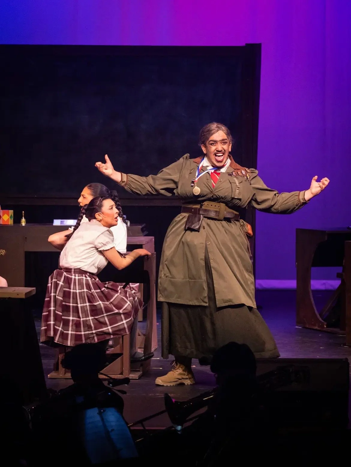 Theater scene with a person in a military costume performing as a soldier and two women in school uniforms at a desk, on stage with purple background.
