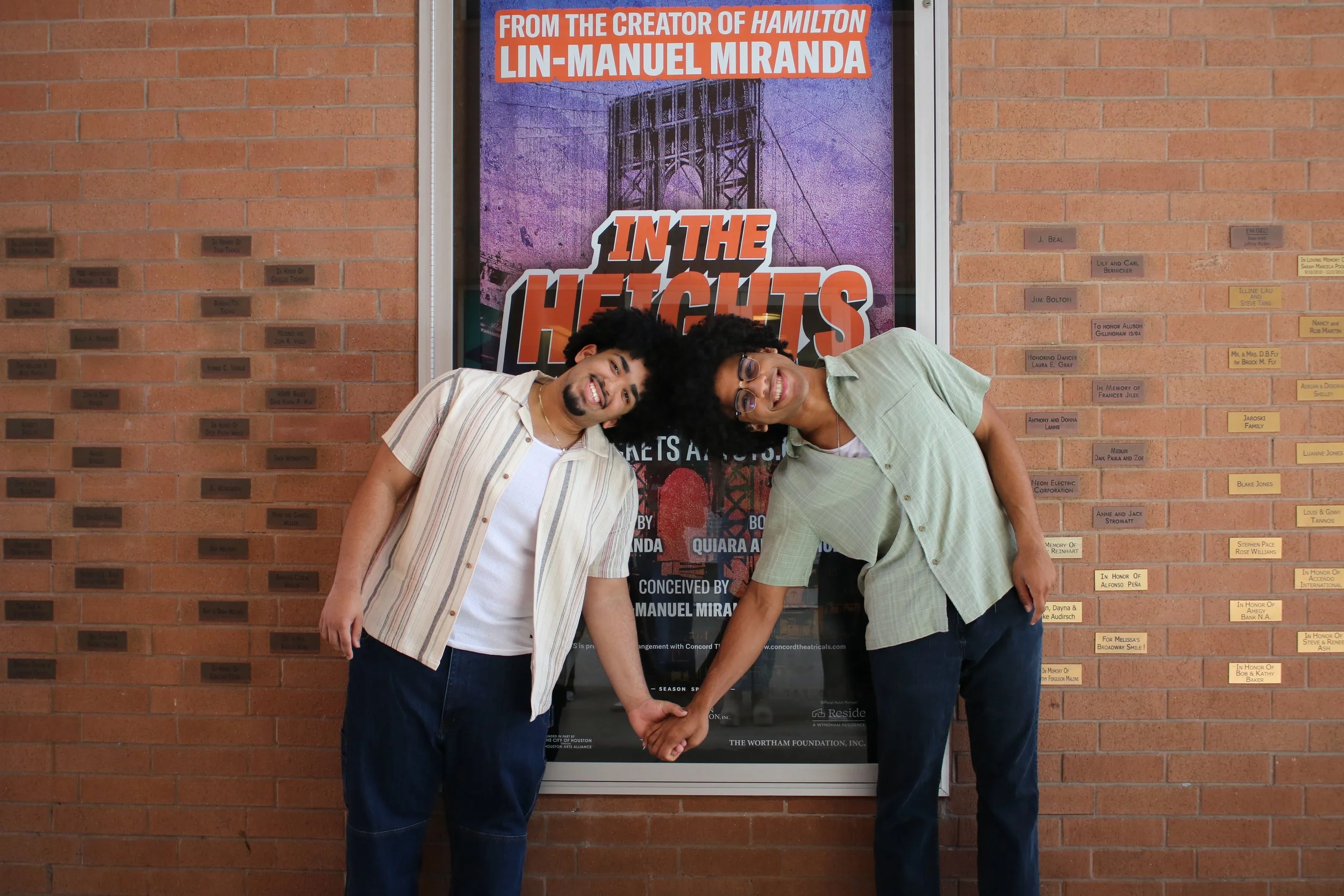 Two young men with curly hair and glasses smiling and holding hands in front of a poster for the musical 'In the Heights,' with a brick wall and memorial plaques in the background.