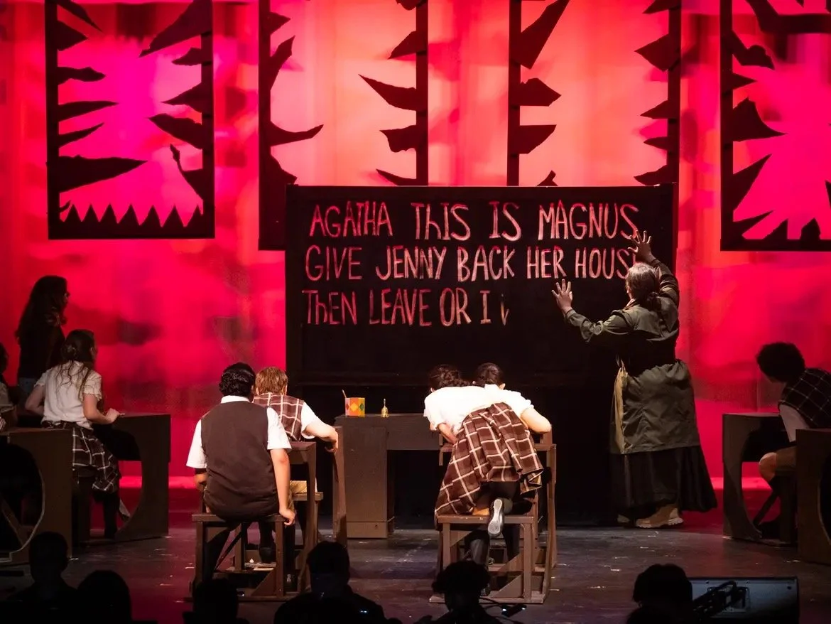 Stage scene from a theatrical play with a blackboard displaying the text 'Agatha this is Magnus. Give Jenny back her house. Then leave or I'. Several actors dressed in school uniforms are on stage, with a woman directing or narrating.