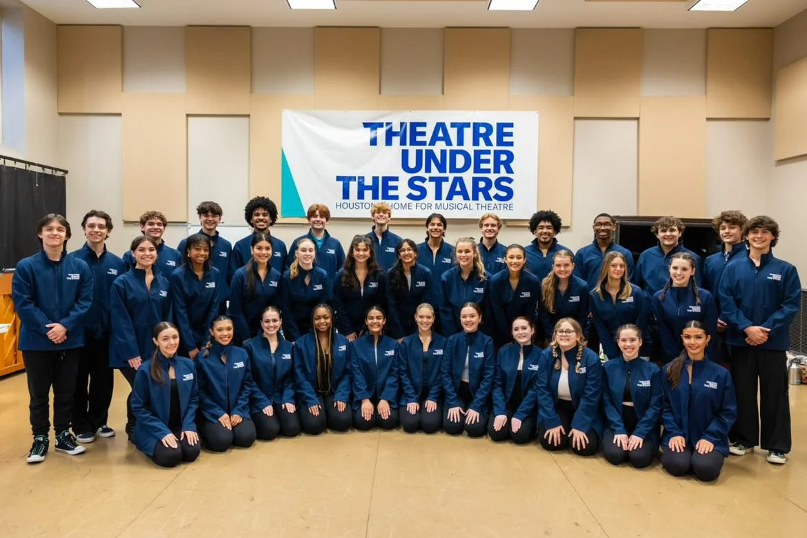 A group of young people in matching blue jackets posed for a photo in a music or theater room. Behind them is a banner reading "THEATRE UNDER THE STARS HOUSTON HOME FOR MUSICAL THEATRE."