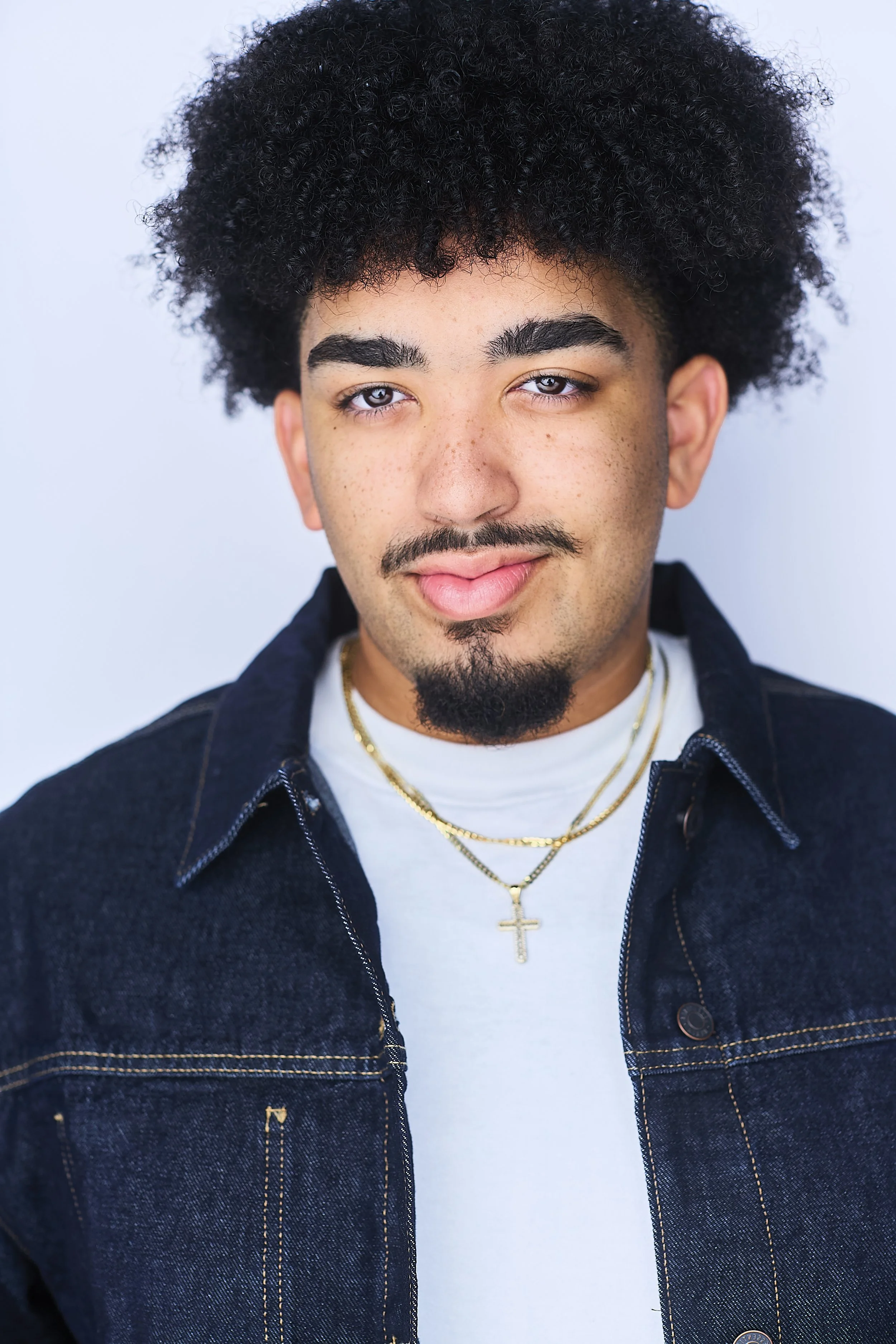 A young man with curly black hair, a mustache, and a goatee, wearing a denim jacket over a white shirt, accessorized with gold necklaces including a cross pendant, looking directly at the camera with a slight smile.