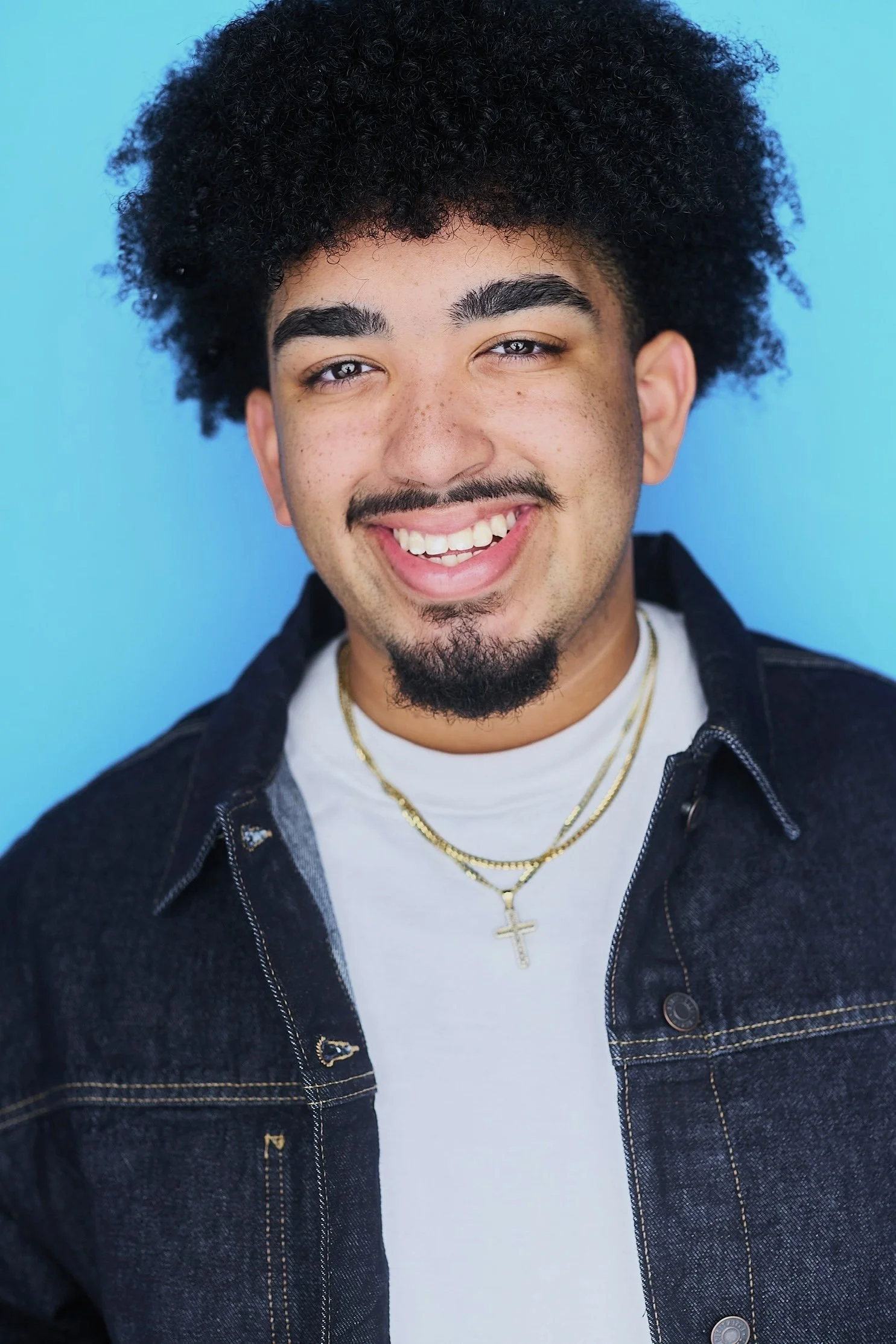 Close-up of a smiling young man with curly black hair, light eyes, freckles, a goatee, wearing layered gold necklaces, a white t-shirt, and a dark denim jacket against a blue background.