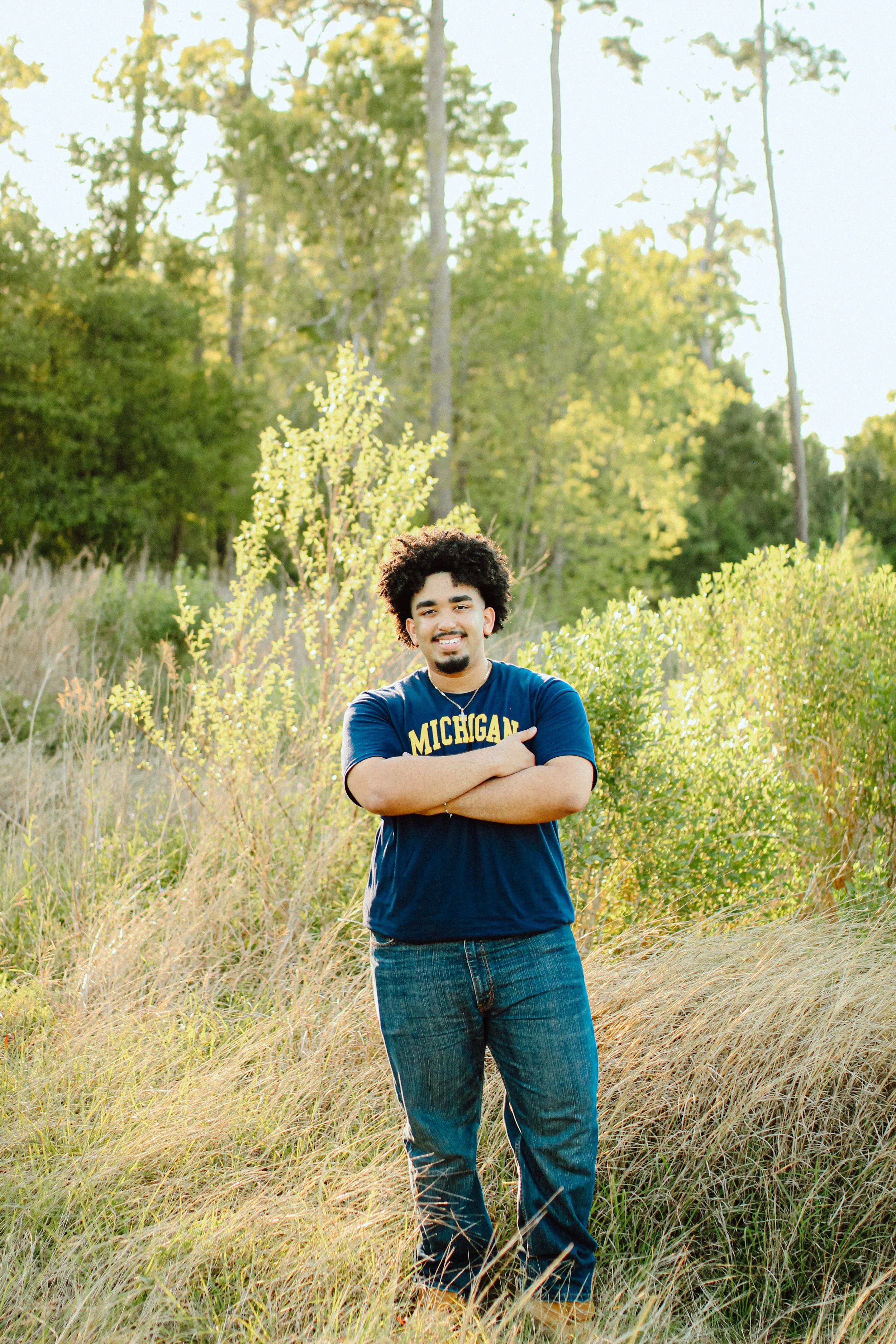 A young man with curly hair and a beard standing outdoors in a grassy field with trees, wearing a blue Michigan T-shirt and jeans, smiling with arms crossed.