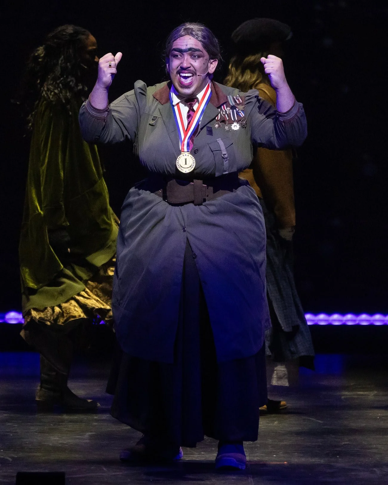 A performer with long hair, wearing a costume with medals, raising fists in a theatrical stage setting with dark background and two other performers behind him.