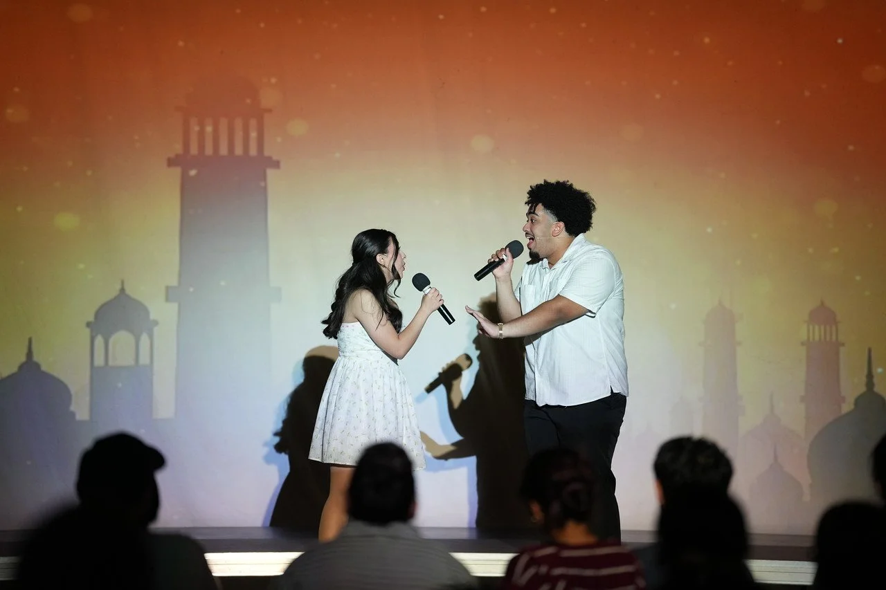 A young woman and a young man are singing into microphones on stage during a performance, with an audience watching. The background features a colorful scene with star-like lights and silhouettes of domed towers or minarets.