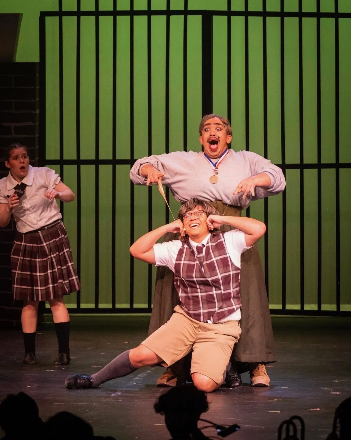 Theater scene with four actors in school uniforms, one wearing a medal, on stage with green and black striped background, performing a comedic scene.