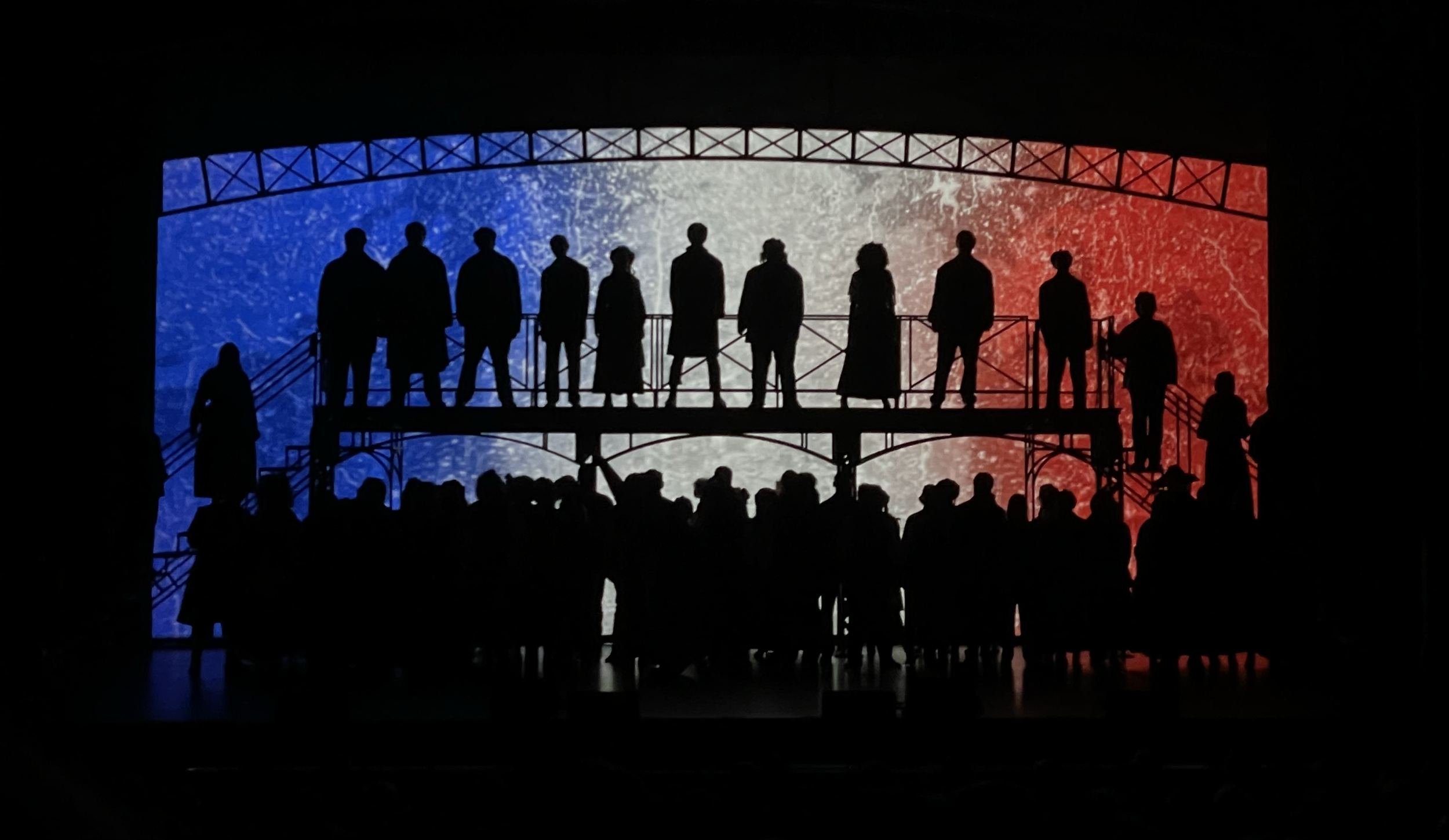 Silhouettes of people on a stage with a colorful blue, white, and red background, resembling the French flag.