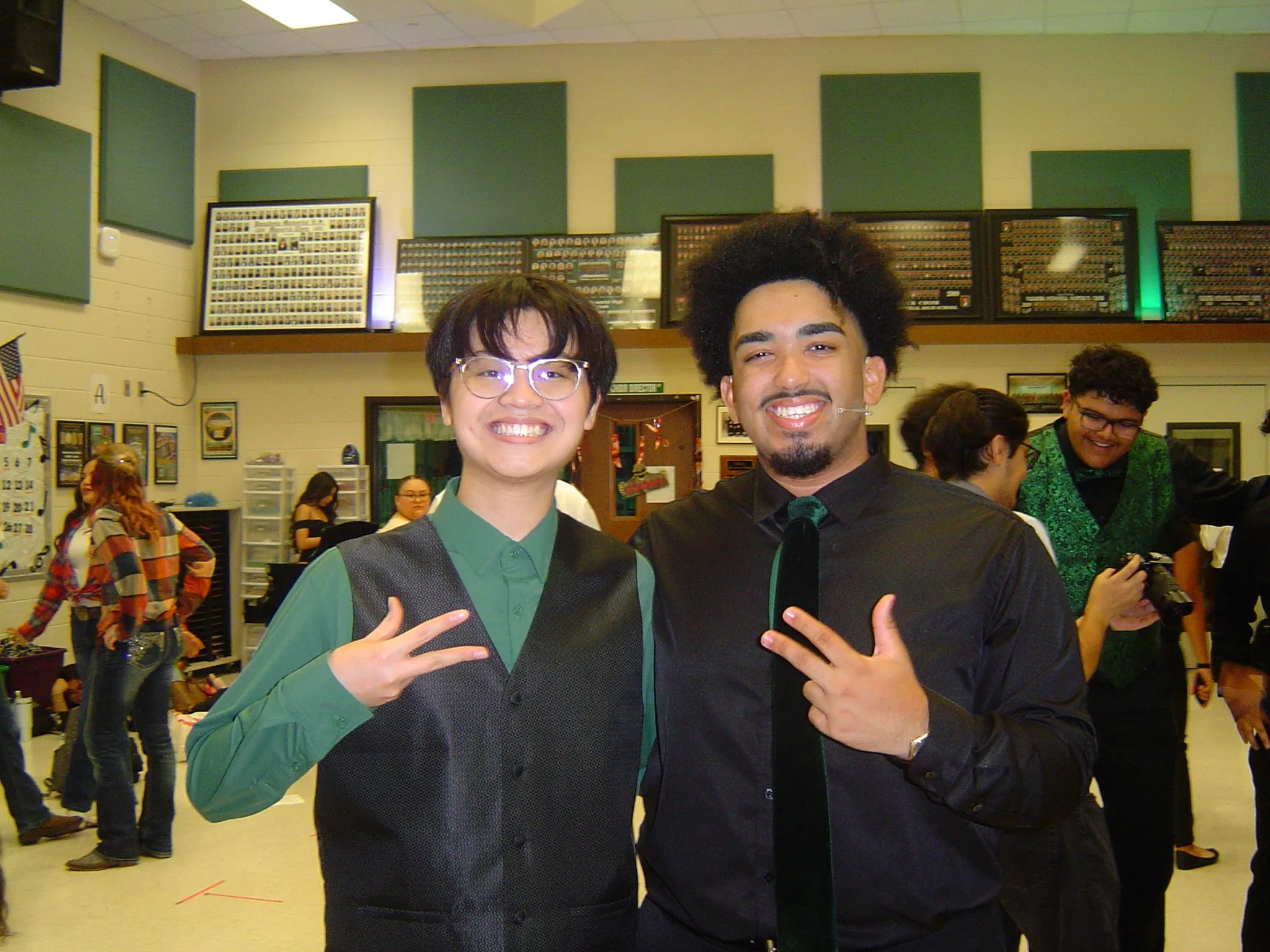 Two young men dressed in traditional graduation gowns and mortarboards happily posing for a photo, making peace signs with their hands, in a celebration environment with other people in the background.
