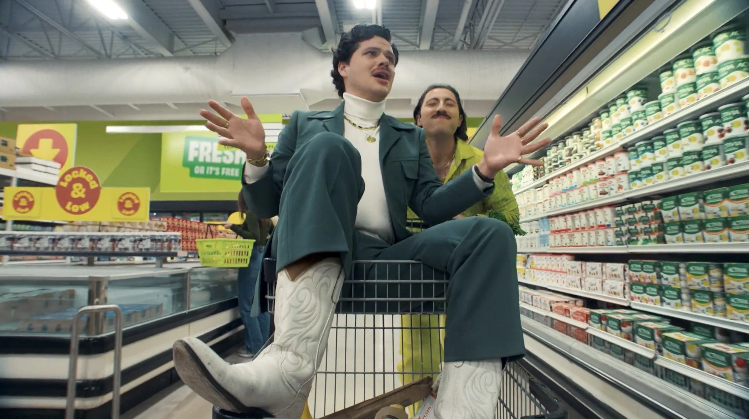 Two men sitting in a shopping cart in a grocery store. The man in front is wearing a teal jacket, white turtleneck, and white cowboy boots. The man behind is wearing a yellow shirt with floral patterns. They are surrounded by shelves of dairy products and other grocery items.