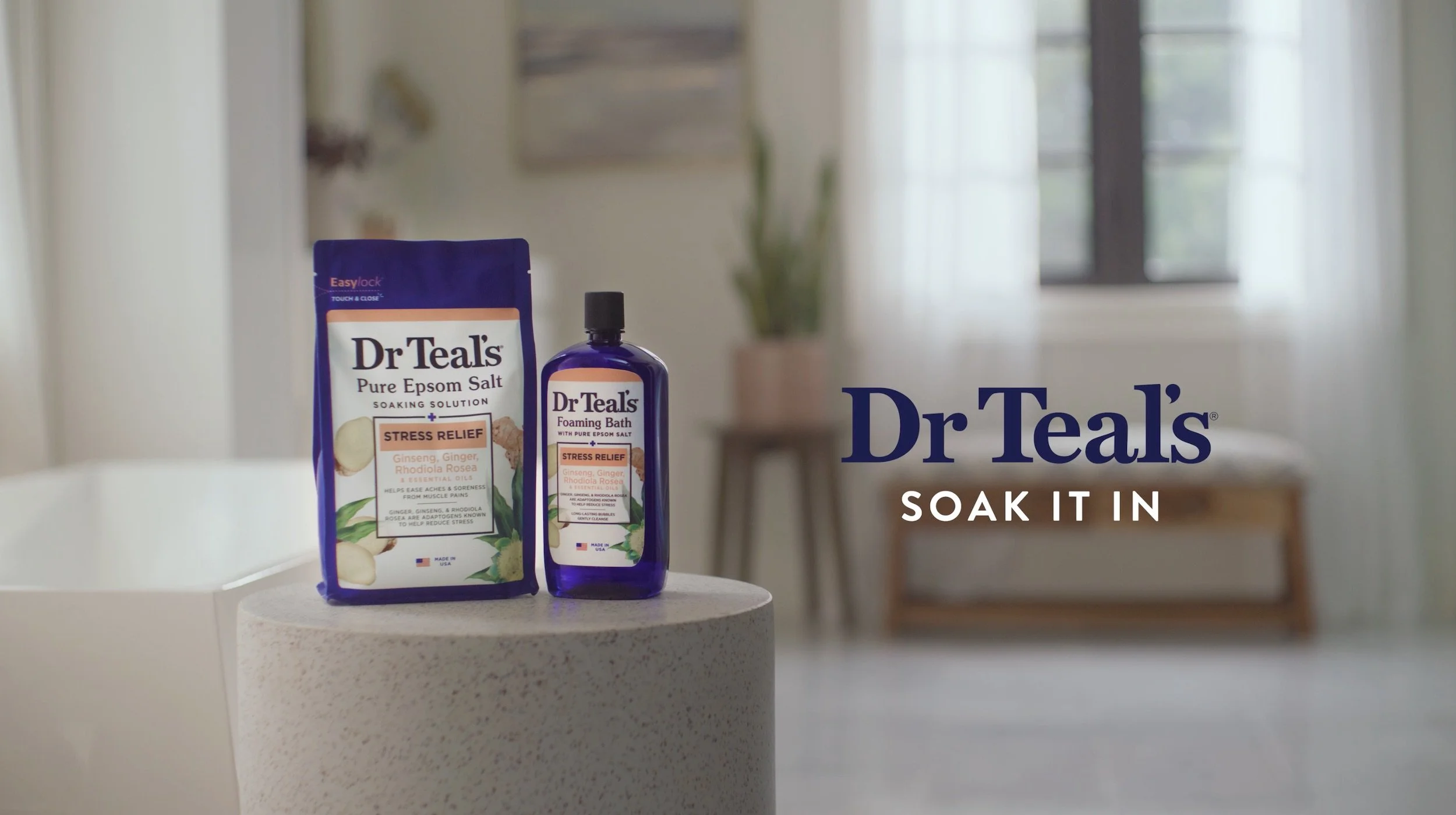 Image of Dr Teal's Stress Relief products, including a bottle of foaming bath and a packet of pure epsom salt, placed on a small white table in a bright room with a window and indoor plants in the background. The text on the image reads 'Dr Teal's SOAK IT IN'.