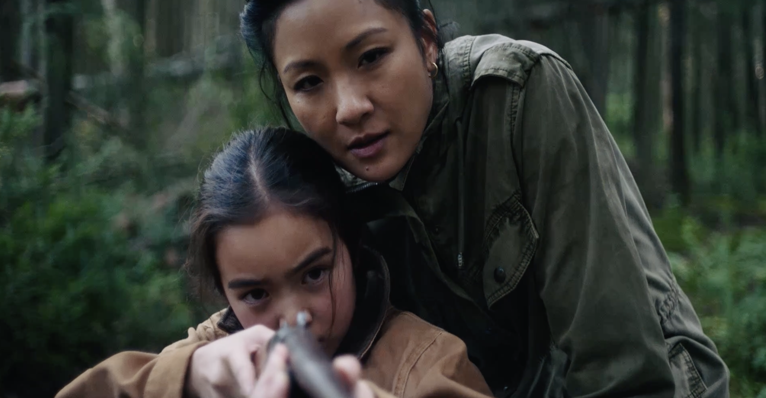 Two women in a forest, one aiming a gun and the other leaning to the side, both with intense expressions.