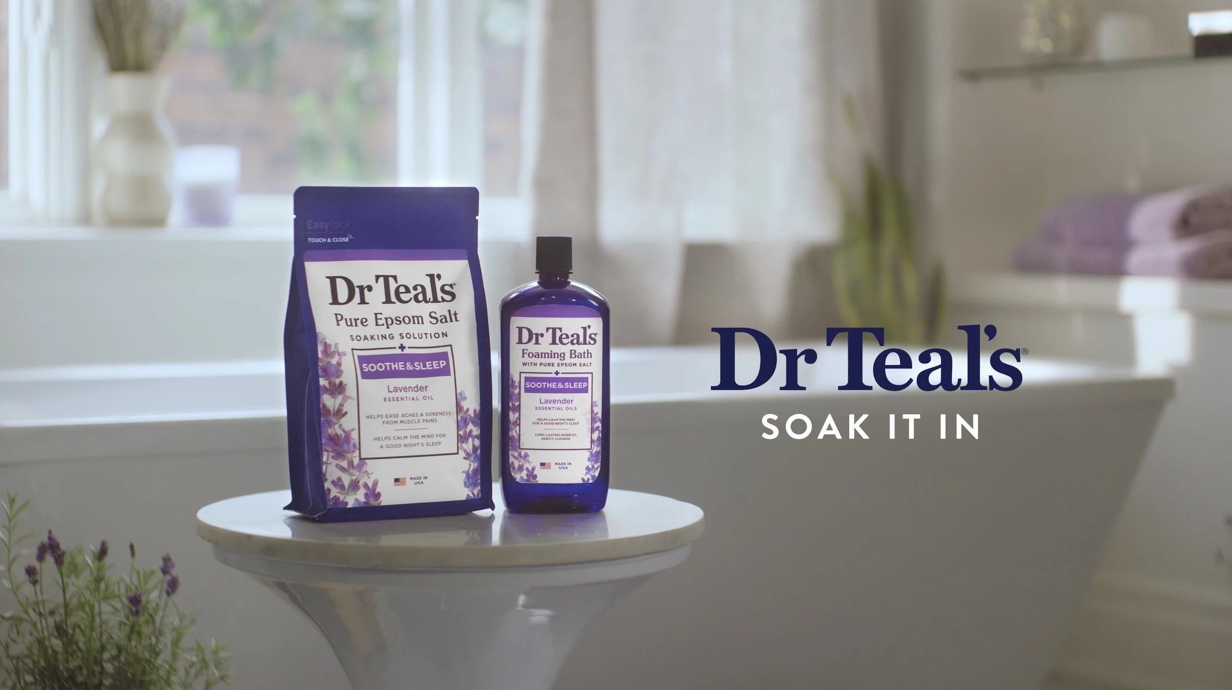 Two lavender scented Dr. Teal's bath products, one bottle of foaming bath and one bag of soak solution, placed on a white side table in front of a window with sheer curtains. The side table has some purple flowers in the foreground, and there is additional purple towels on a surface in the background.