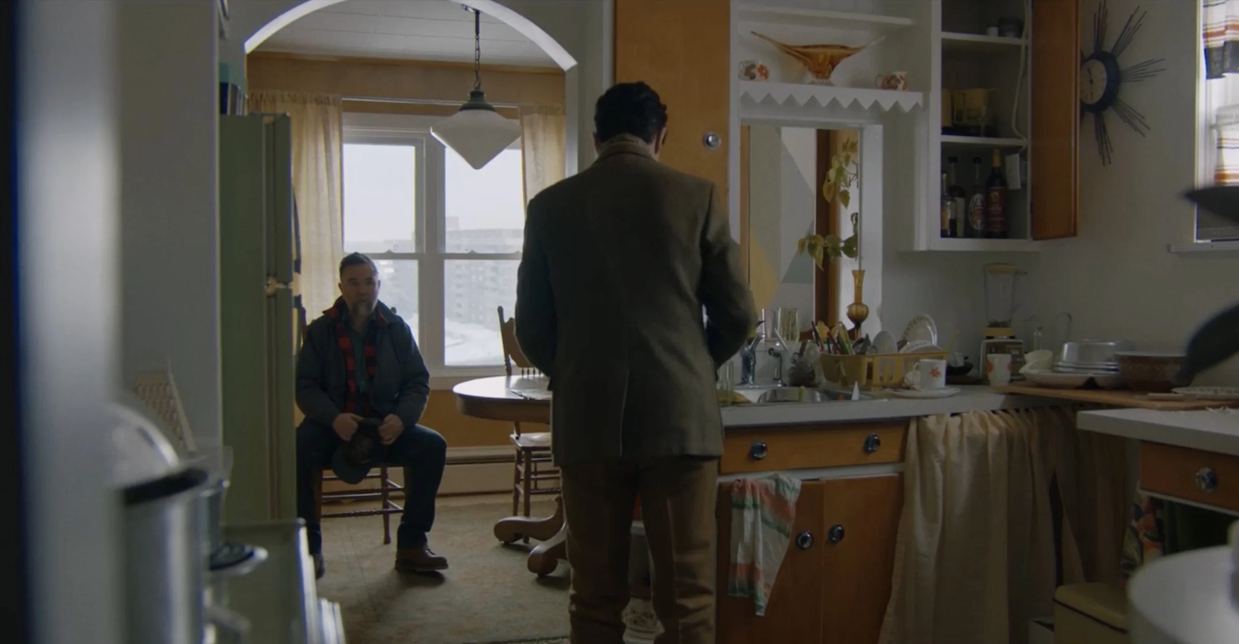 Two men inside a kitchen, one standing with back to the camera and the other sitting on a chair. The kitchen has wooden cabinets, open shelves, and a window with yellow curtains, with various kitchen appliances and items on the counters.