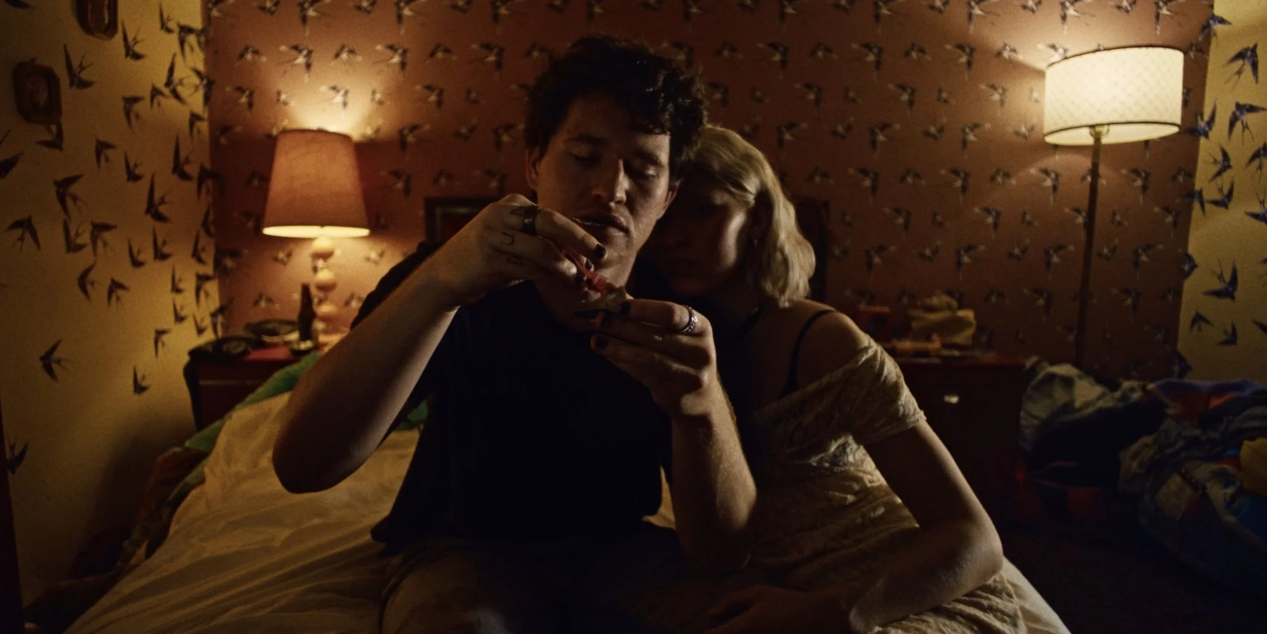 A couple sit on a bed while rolling a joint