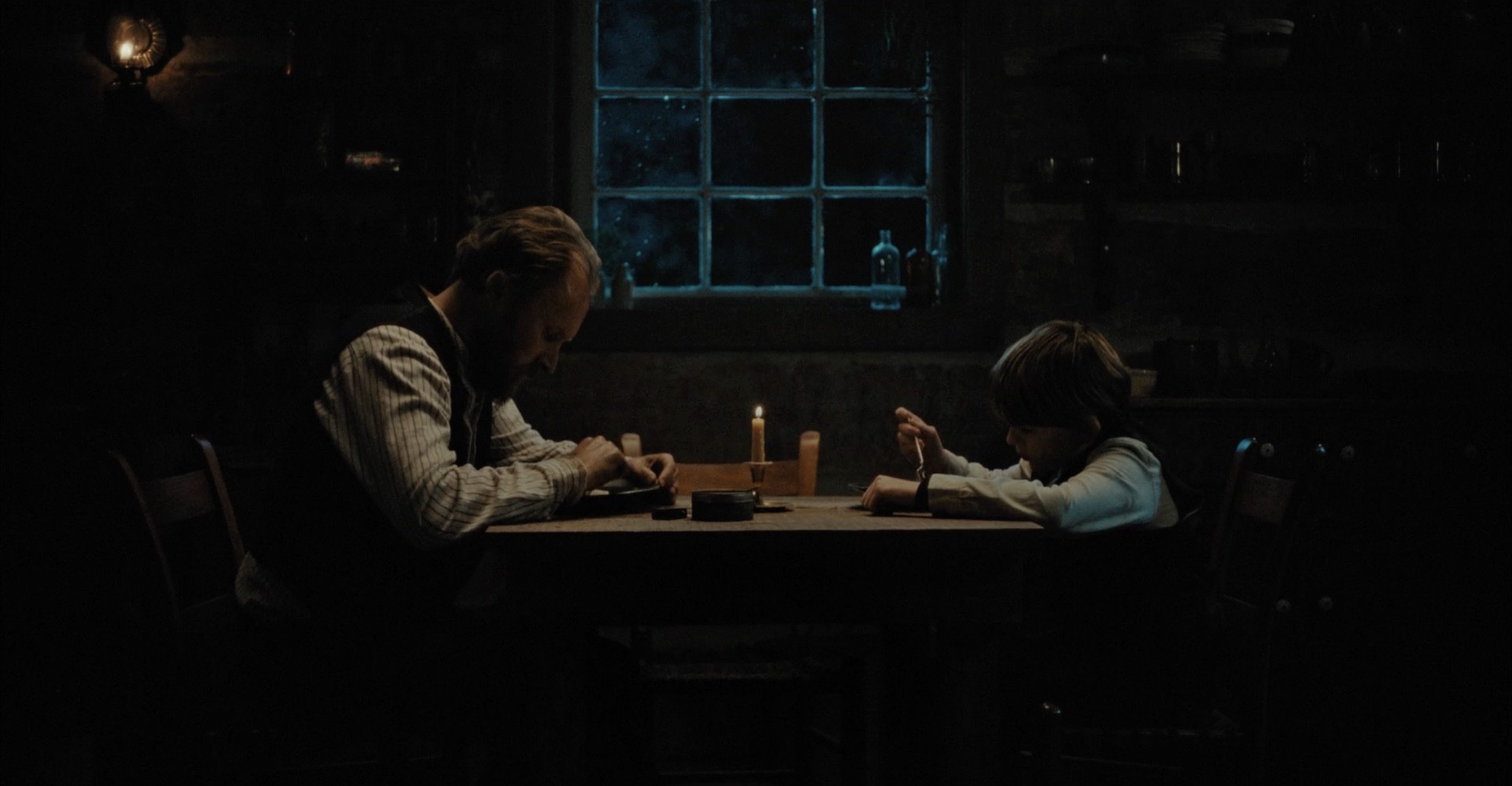 A man and a boy sitting at a wooden table in a dimly lit room, illuminated by a candle, with a window behind them.