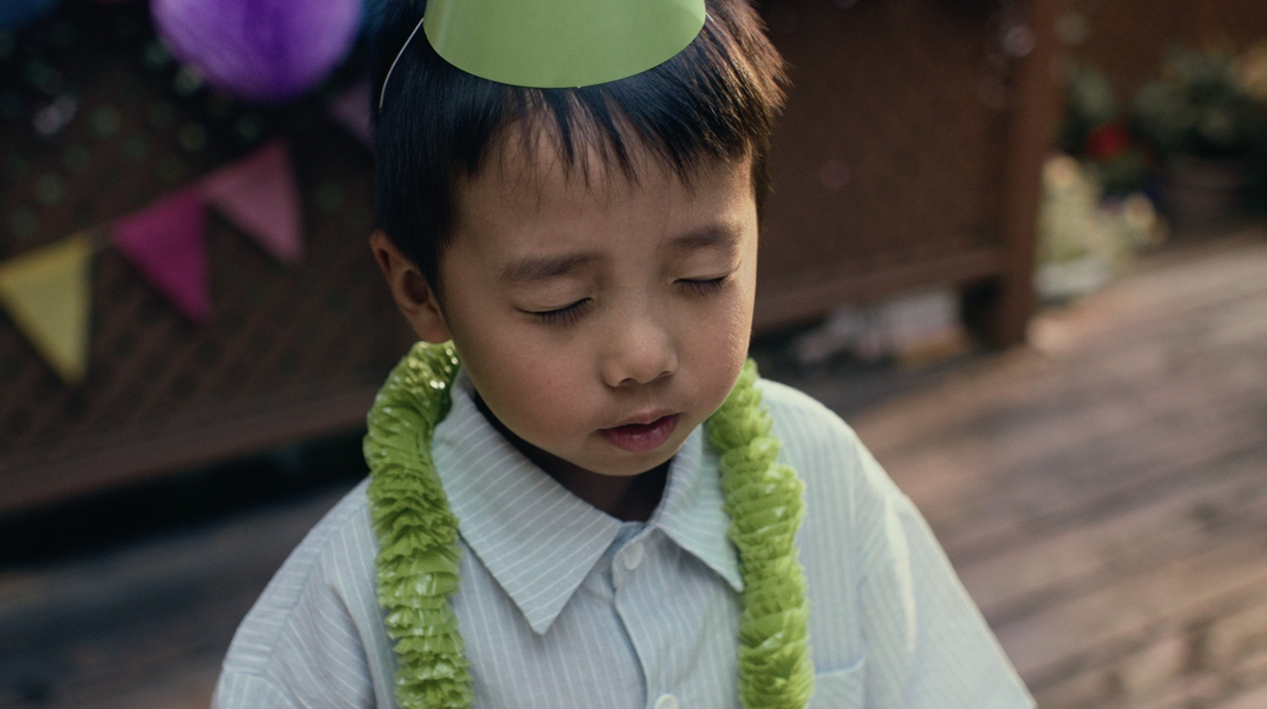 A young boy with closed eyes wearing a light green paper party hat and a bright green lei around his neck, standing outdoors with a blurred background.