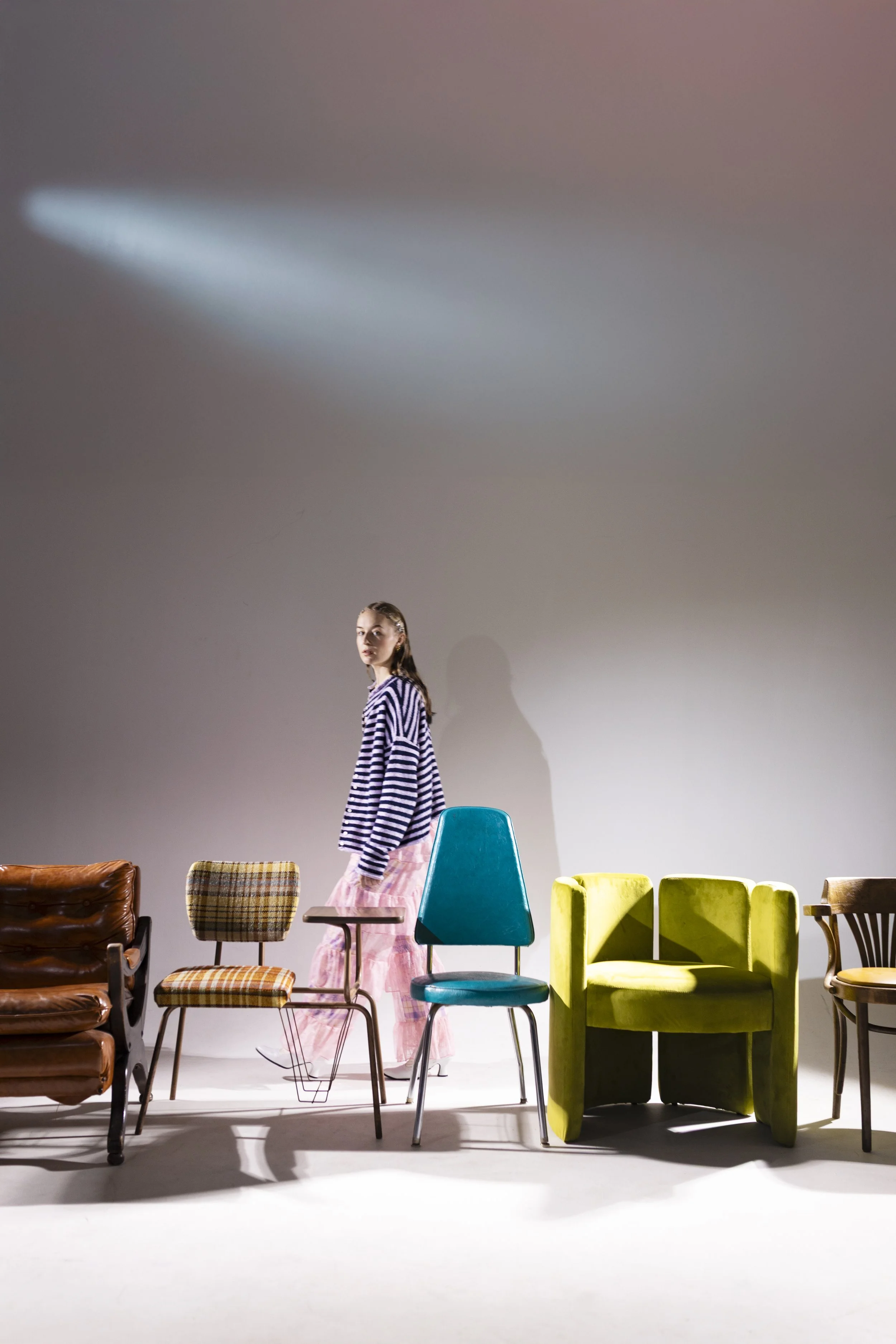 A woman in a striped shirt and pink skirt walking past a row of colorful chairs against a plain gray wall, with a spotlight shadow on the wall
