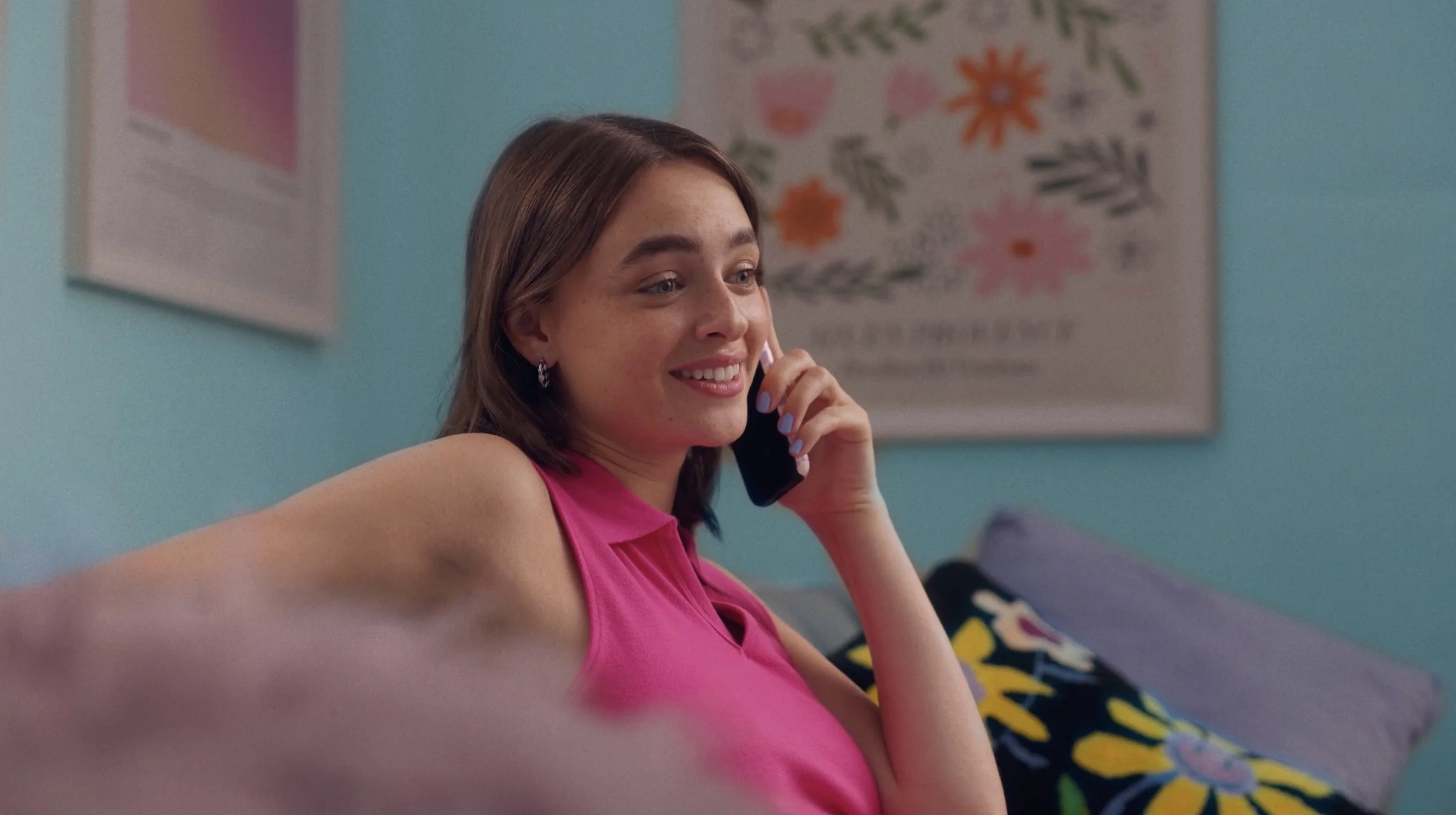 A young woman with light brown hair and freckles, wearing a pink top, smiling while talking on a black cordless phone in a room with light blue walls and colorful floral and abstract art posters.