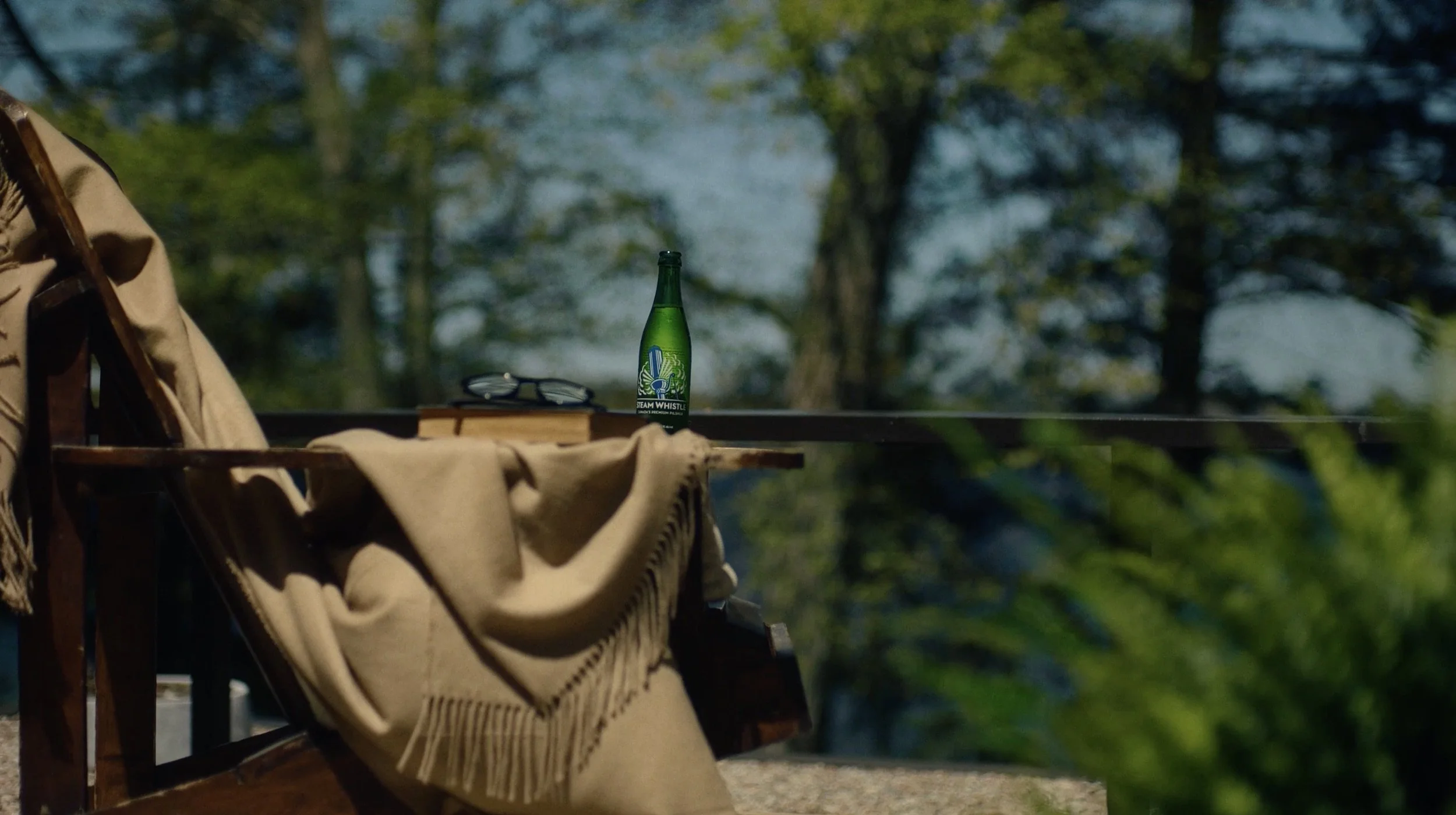 A green glass bottle of Steam Whistle beer on a wooden surface with a pair of sunglasses and a beige blanket draped over a chair, outdoors with trees in the background.