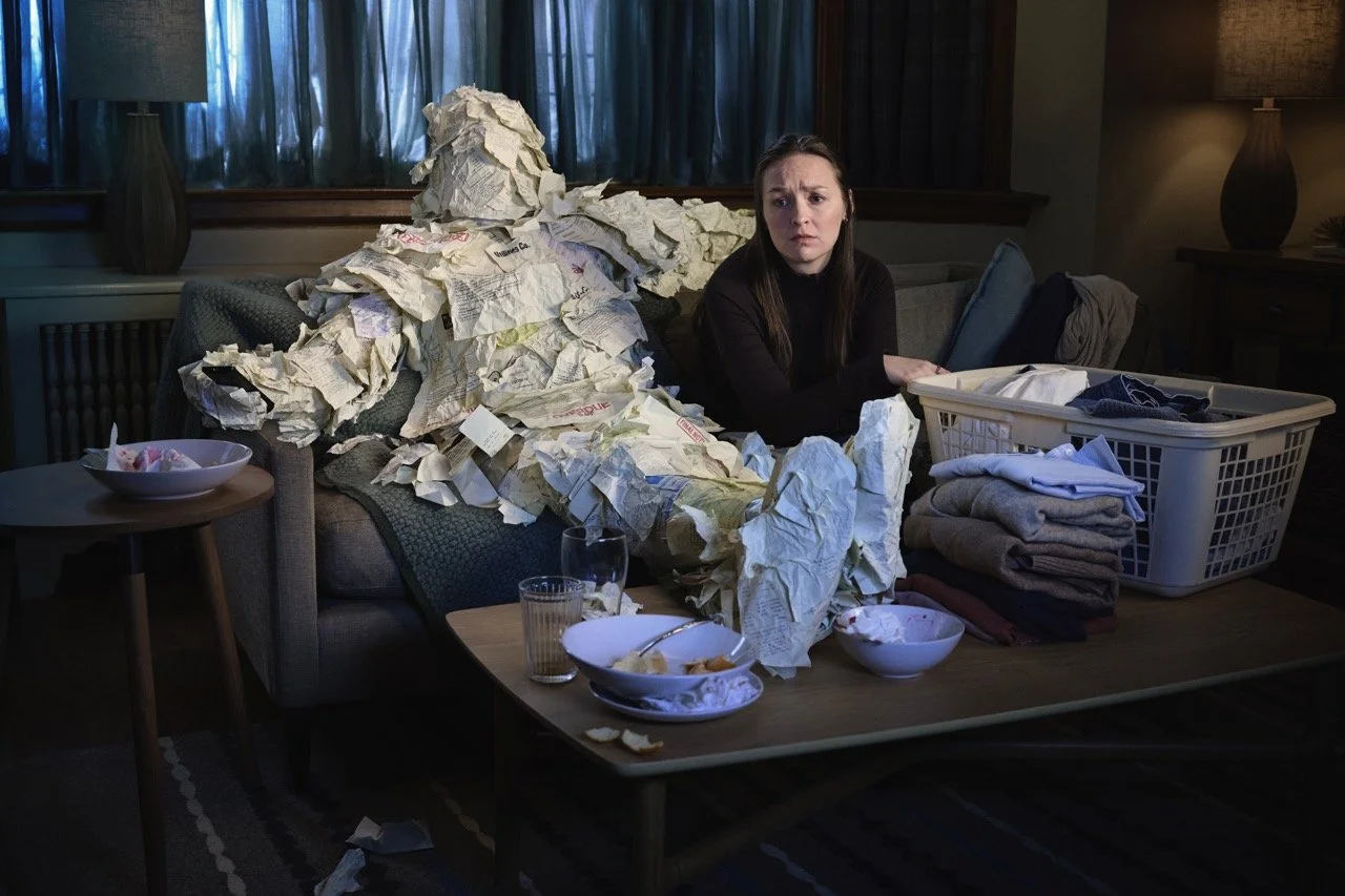 A worried woman sits on a couch with a person 100% covered in bills etc.