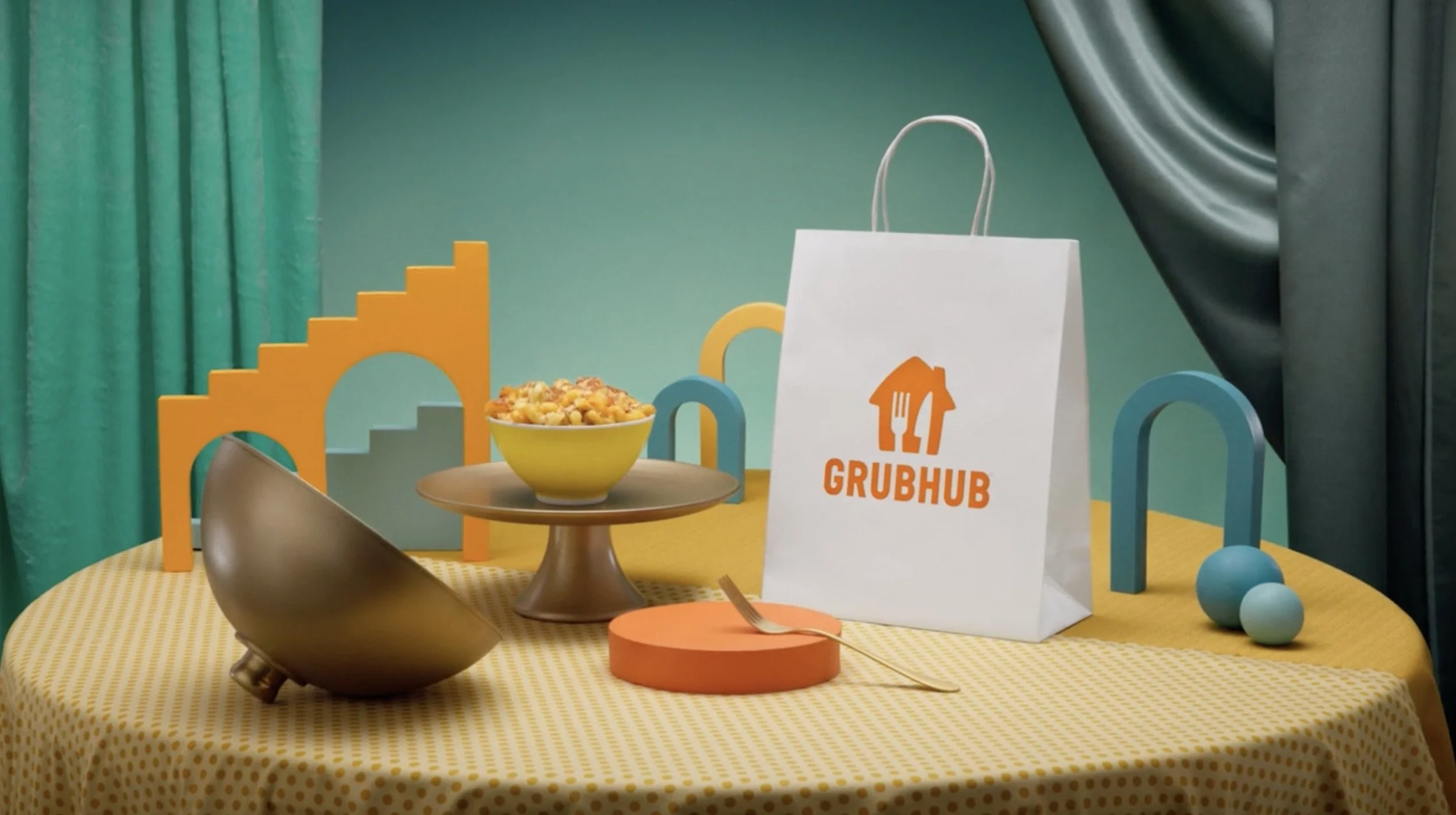 Colorful dining setup with yellow polka dot tablecloth, a bowl of pasta on a gold-colored cake stand, a Grubhub paper bag, and colorful geometric shapes and chairs in the background.