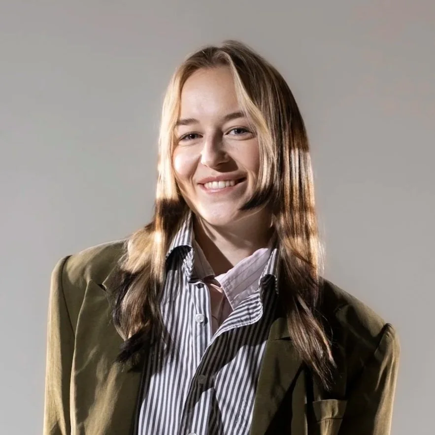Young woman with light brown hair smiling, wearing a striped shirt and olive green jacket against a plain background.