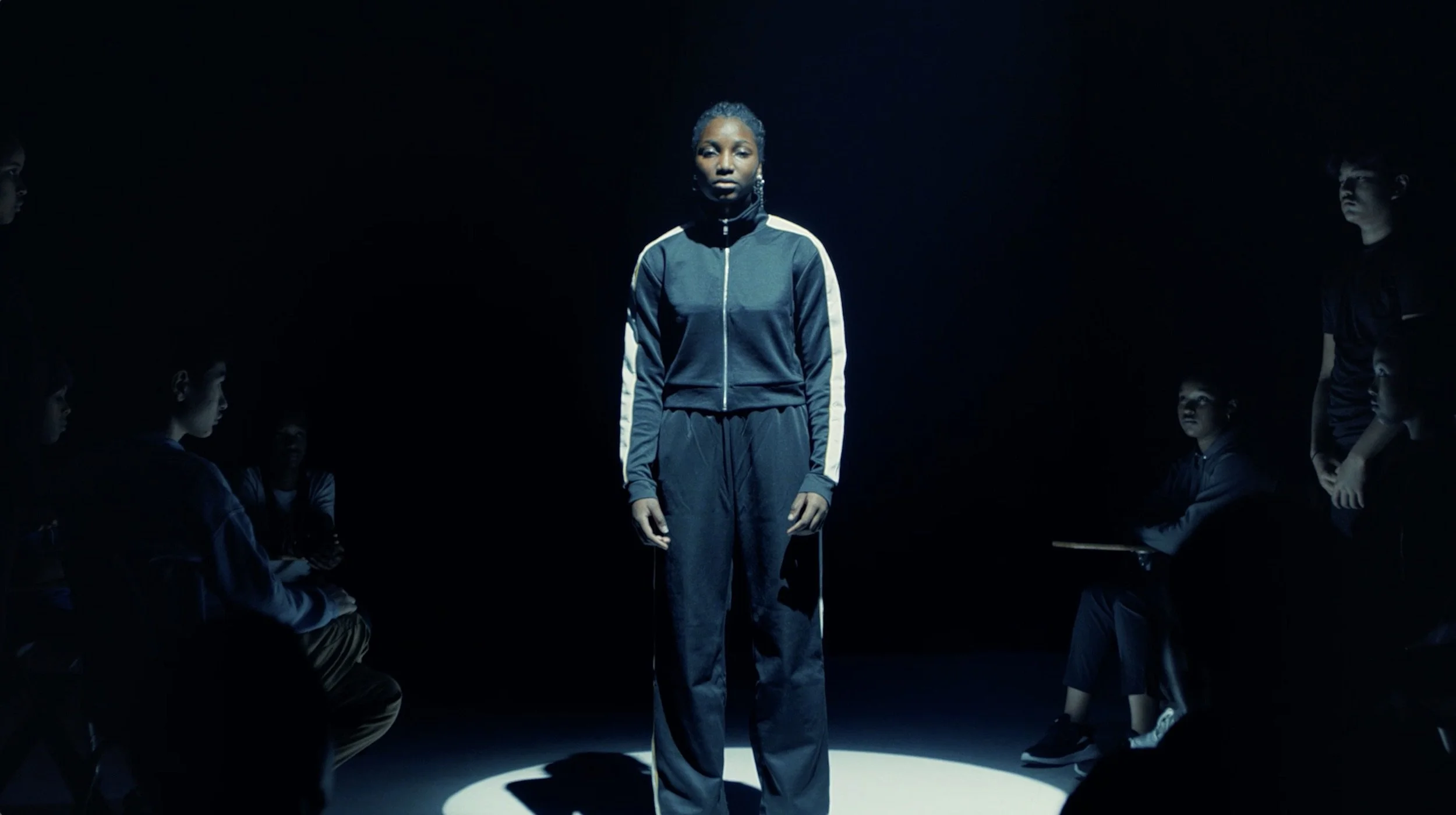 A woman stands in the center of a dark room, surrounded by children sitting and standing around her, illuminated by a spotlight.