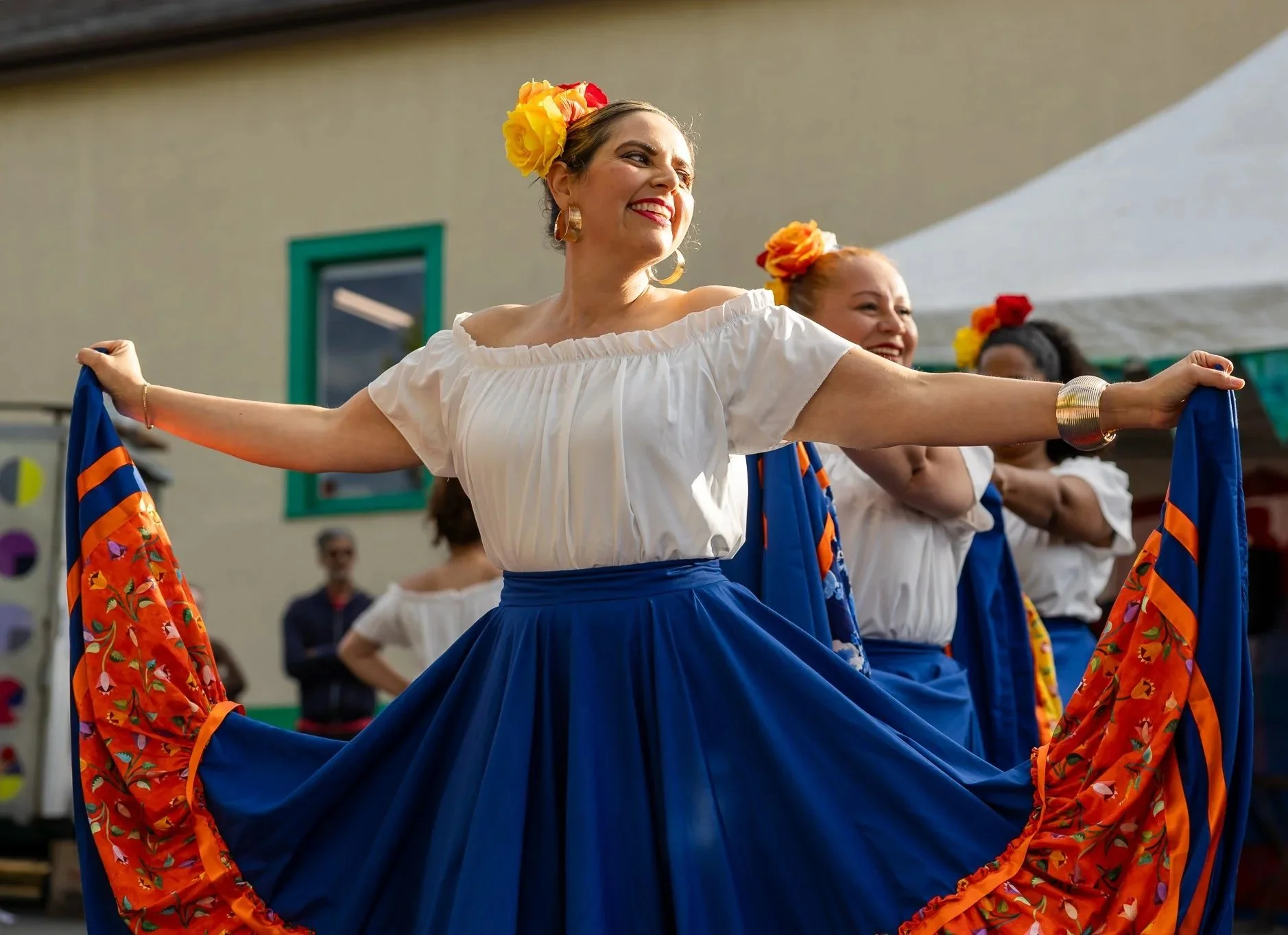 Women in traditional Mexican attire dancing in a group, smiling, wearing colorful floral headpieces and bright skirts.
